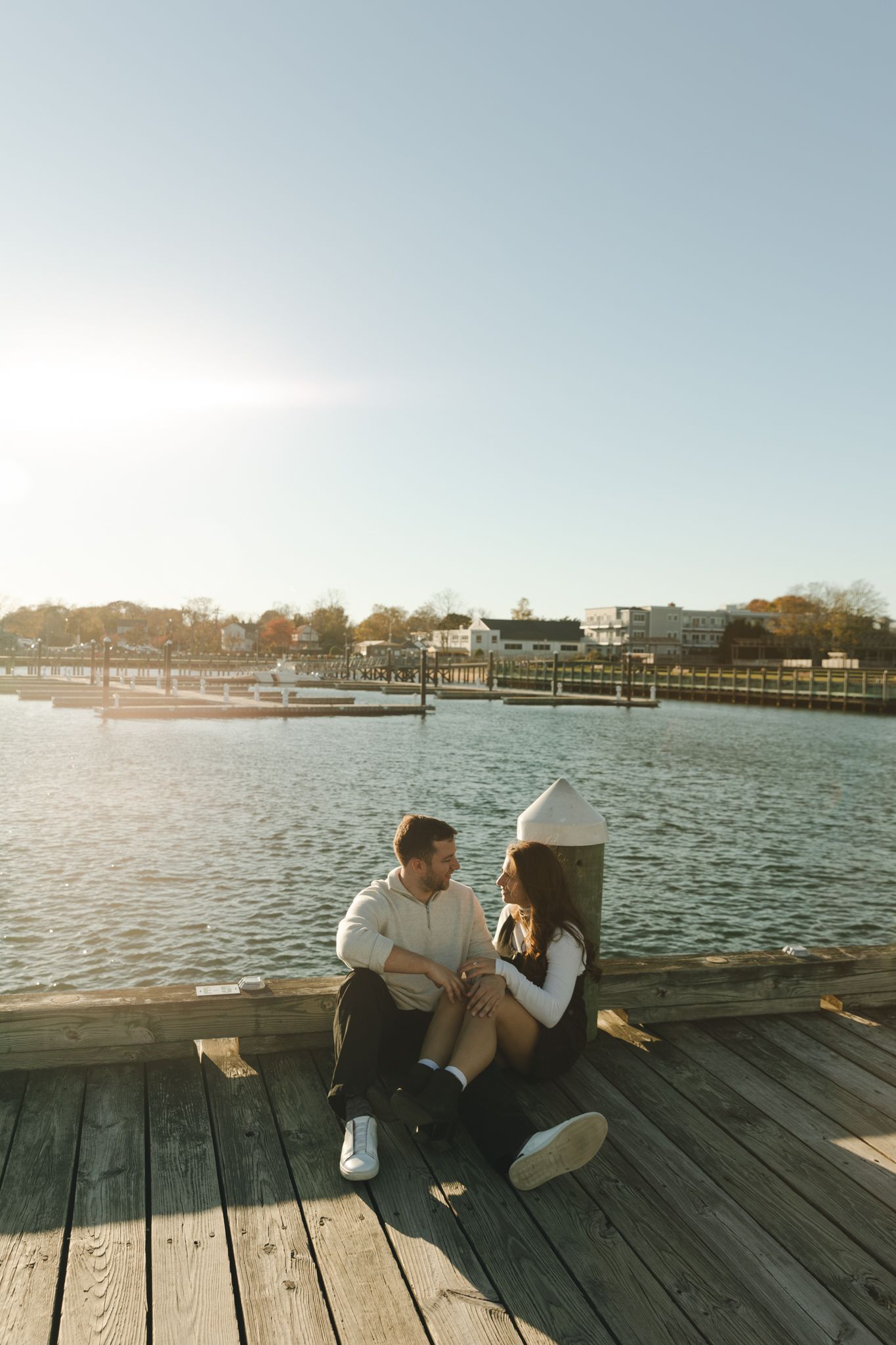 Two people sitting on a wooden pier overlooking a calm harbor at sunset.