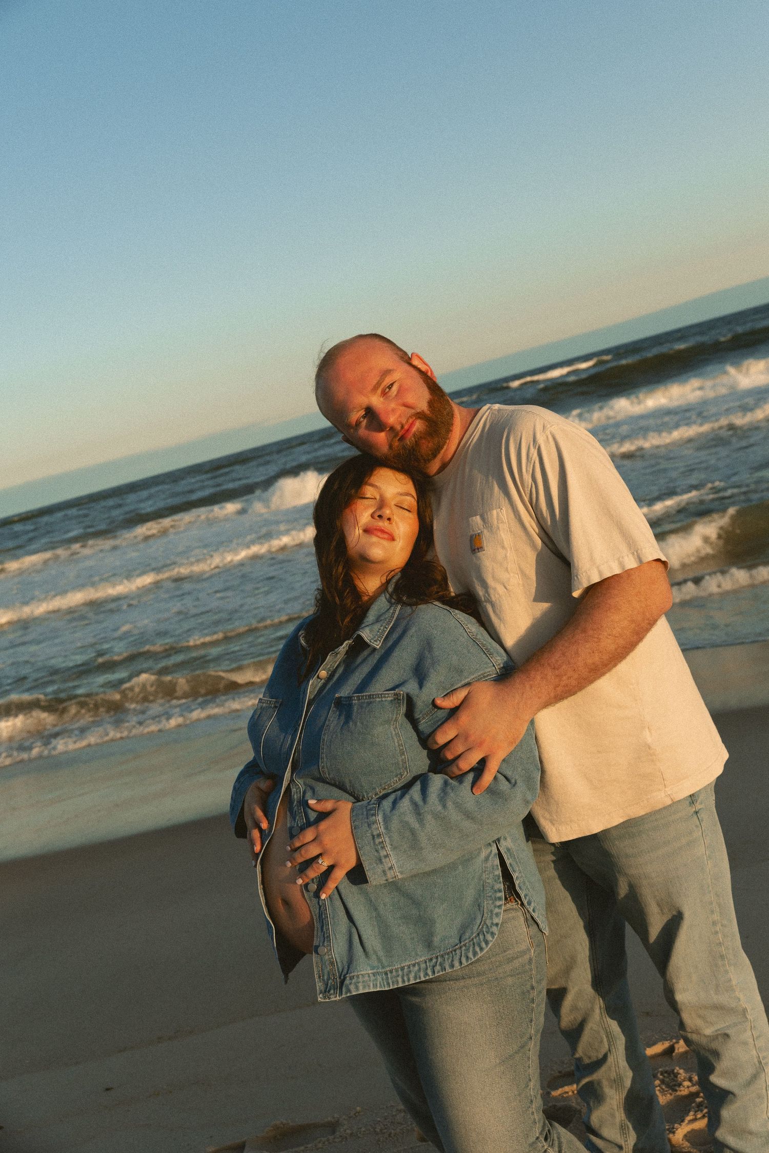 A couple embraces on a beach at sunset, the woman visibly pregnant, waves behind them.