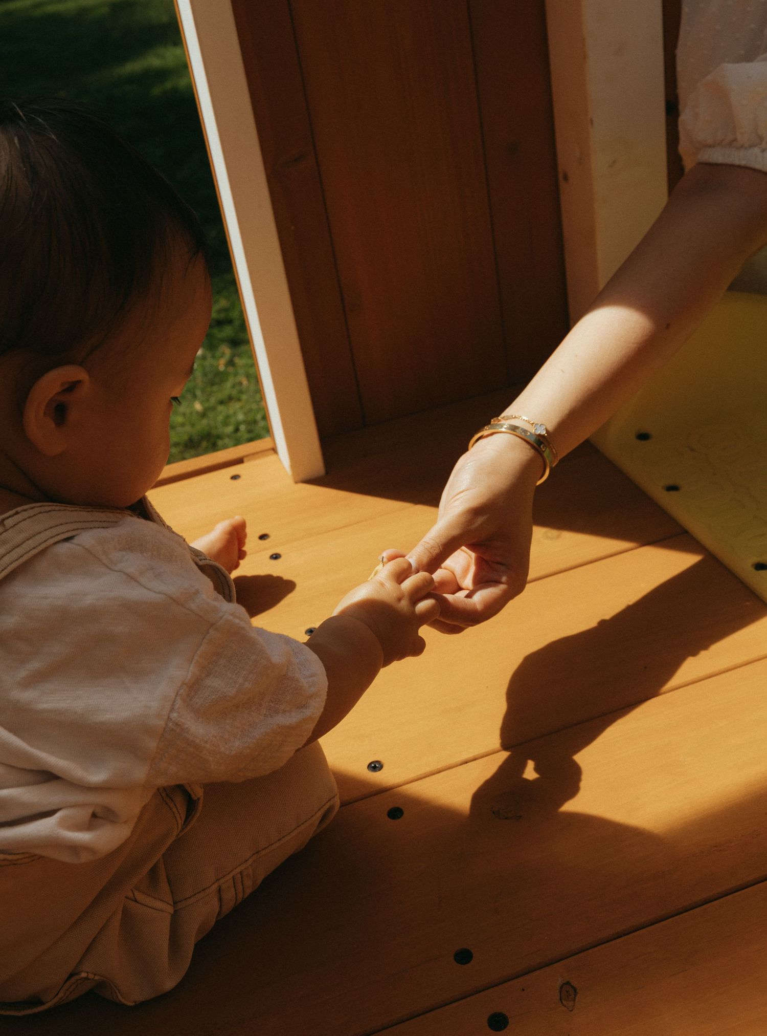 A toddler reaches out to hold an adult's hand on a sunny wooden deck near a playhouse.