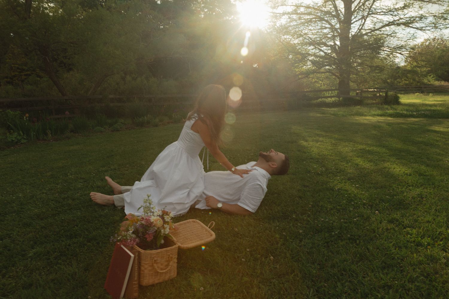 Couple in white attire enjoying a romantic sunset picnic on lush green grass in a sunlit park.