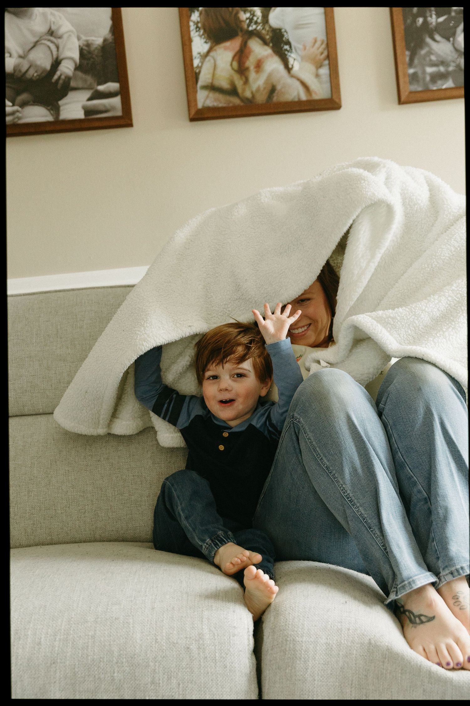 A woman and young child playfully hide under a cozy white blanket together on a gray sofa at home.