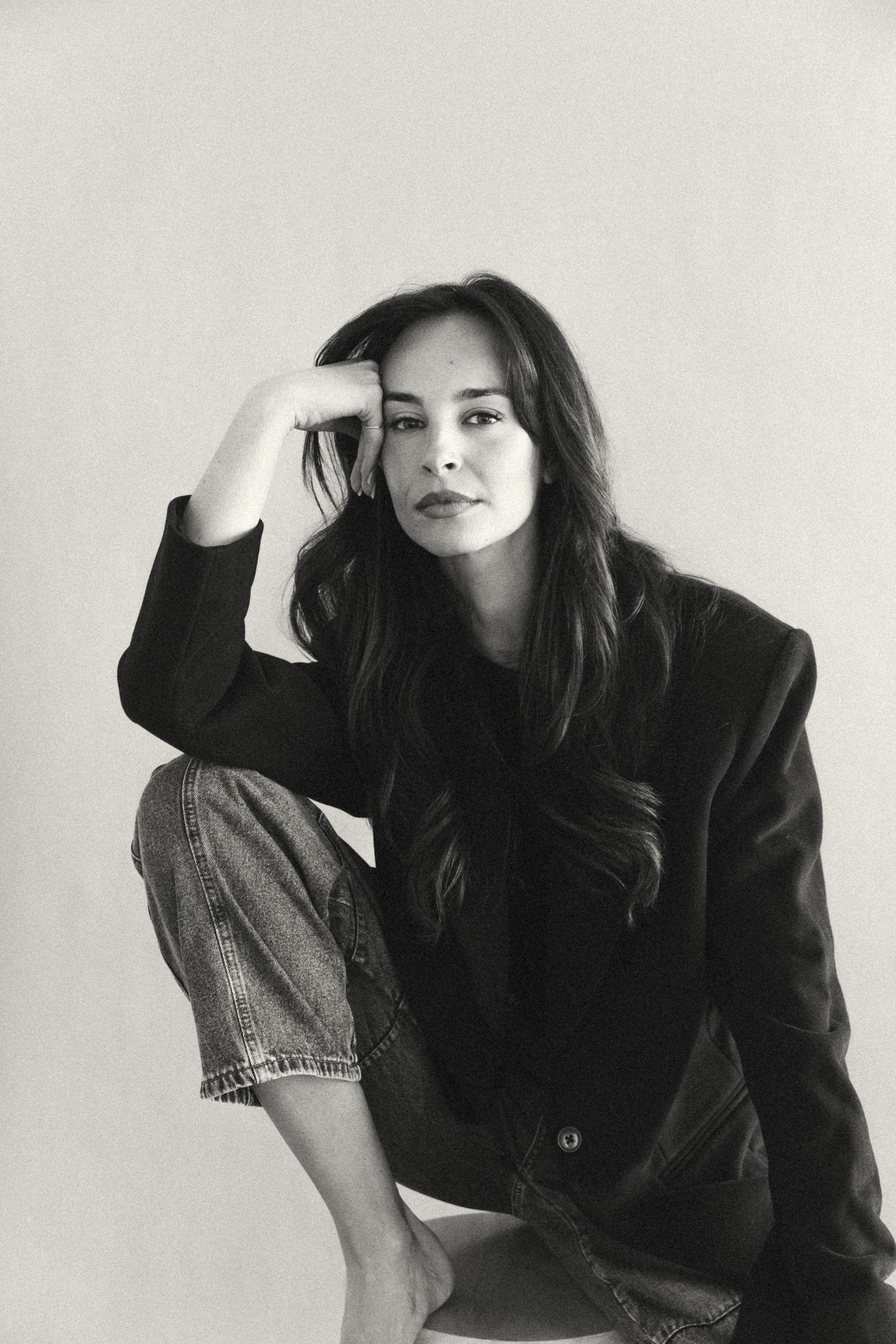 Black and white portrait of a young woman in blazer and jeans sitting relaxed with hand raised to her head.