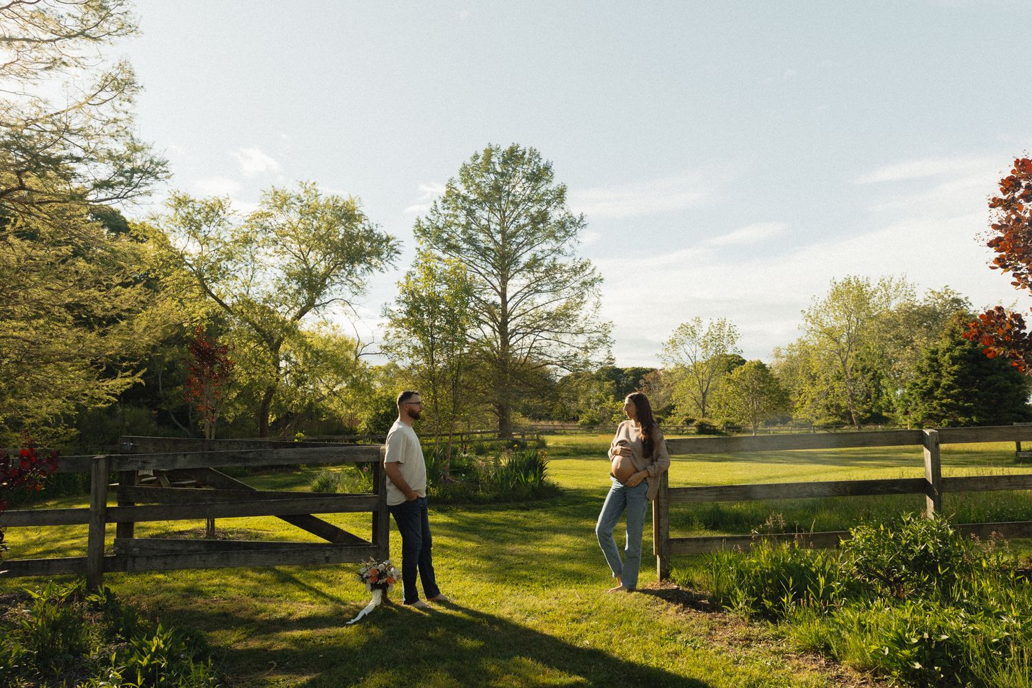 Couple standing on lush green lawn by wooden fence surrounded by trees on sunny spring day.