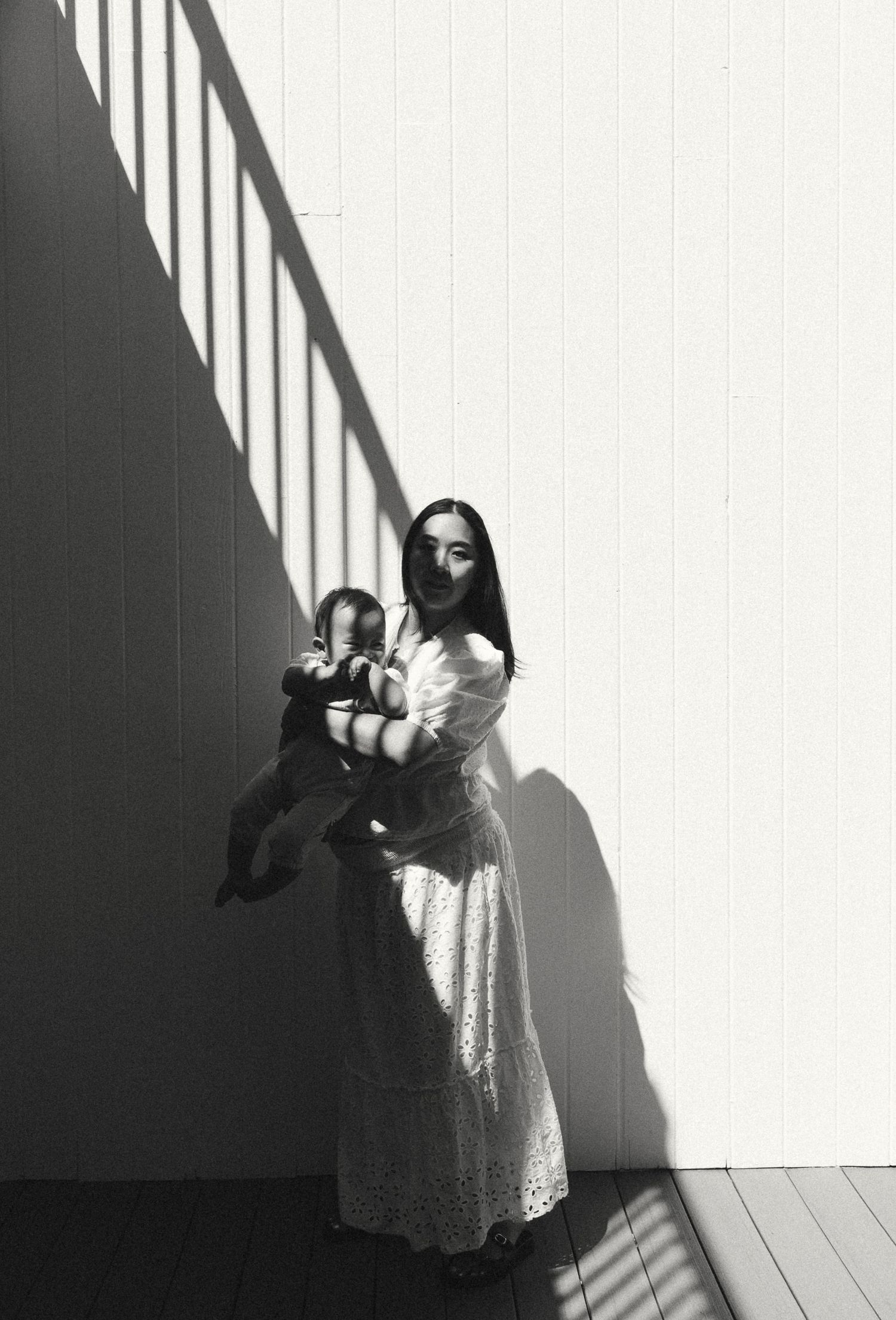 Black and white portrait of mother in white lace dress holding baby against sunlit wall with shadows.
