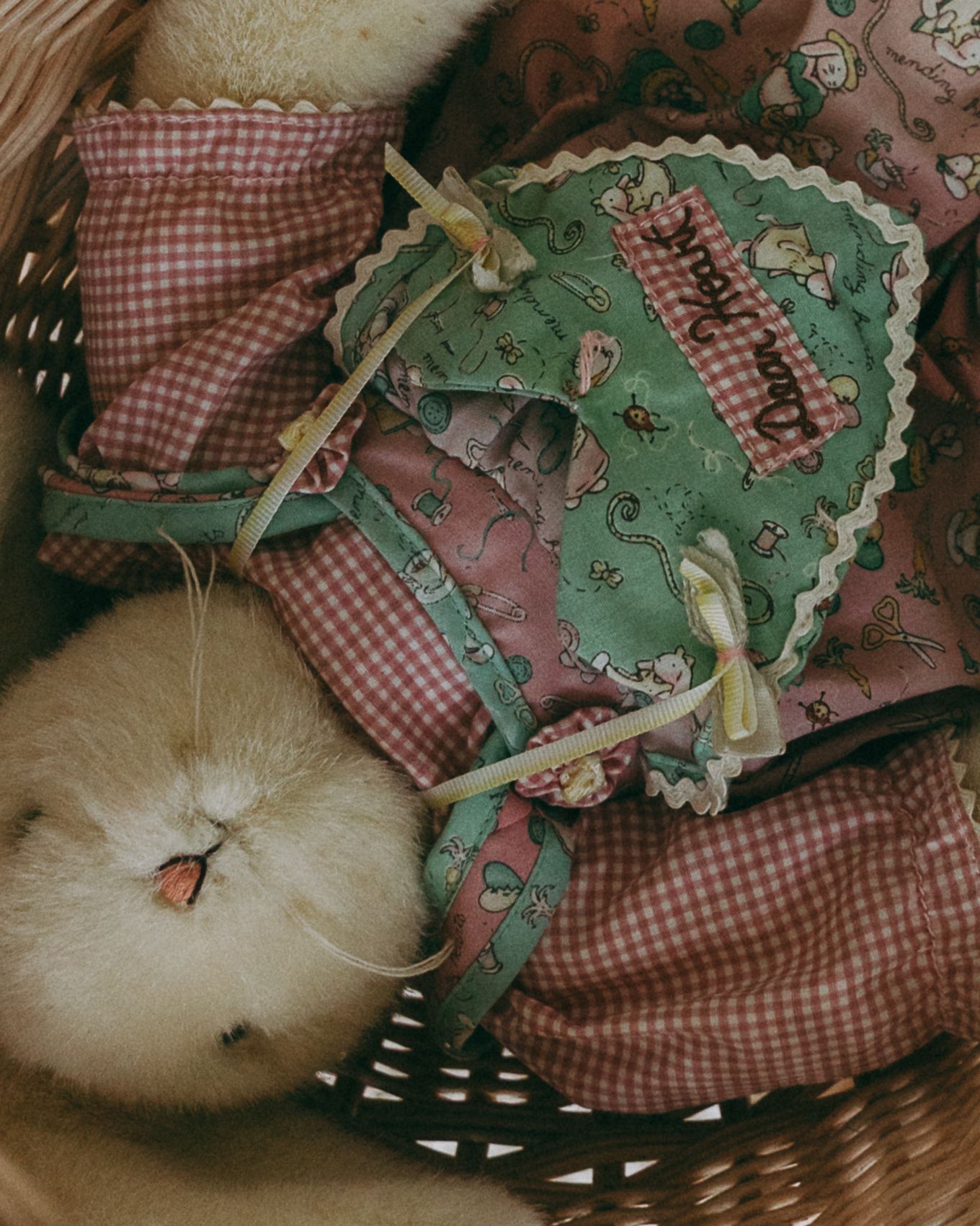A baby chick rests in a wicker basket surrounded by pink gingham fabric and a green floral quilted heart decoration.