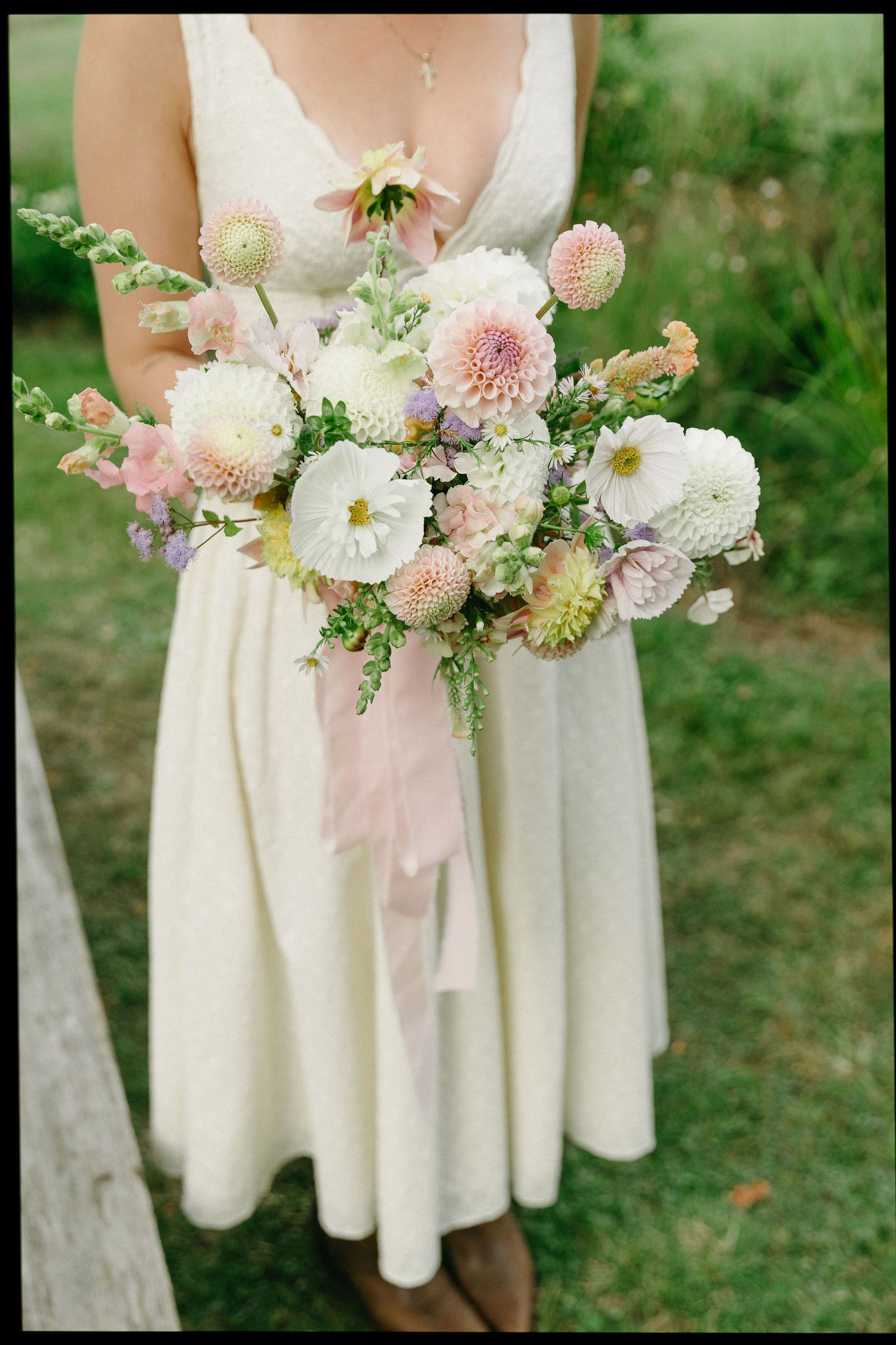 A bride in a white dress holds a lush bouquet of dahlias and wildflowers tied with a soft pink ribbon.