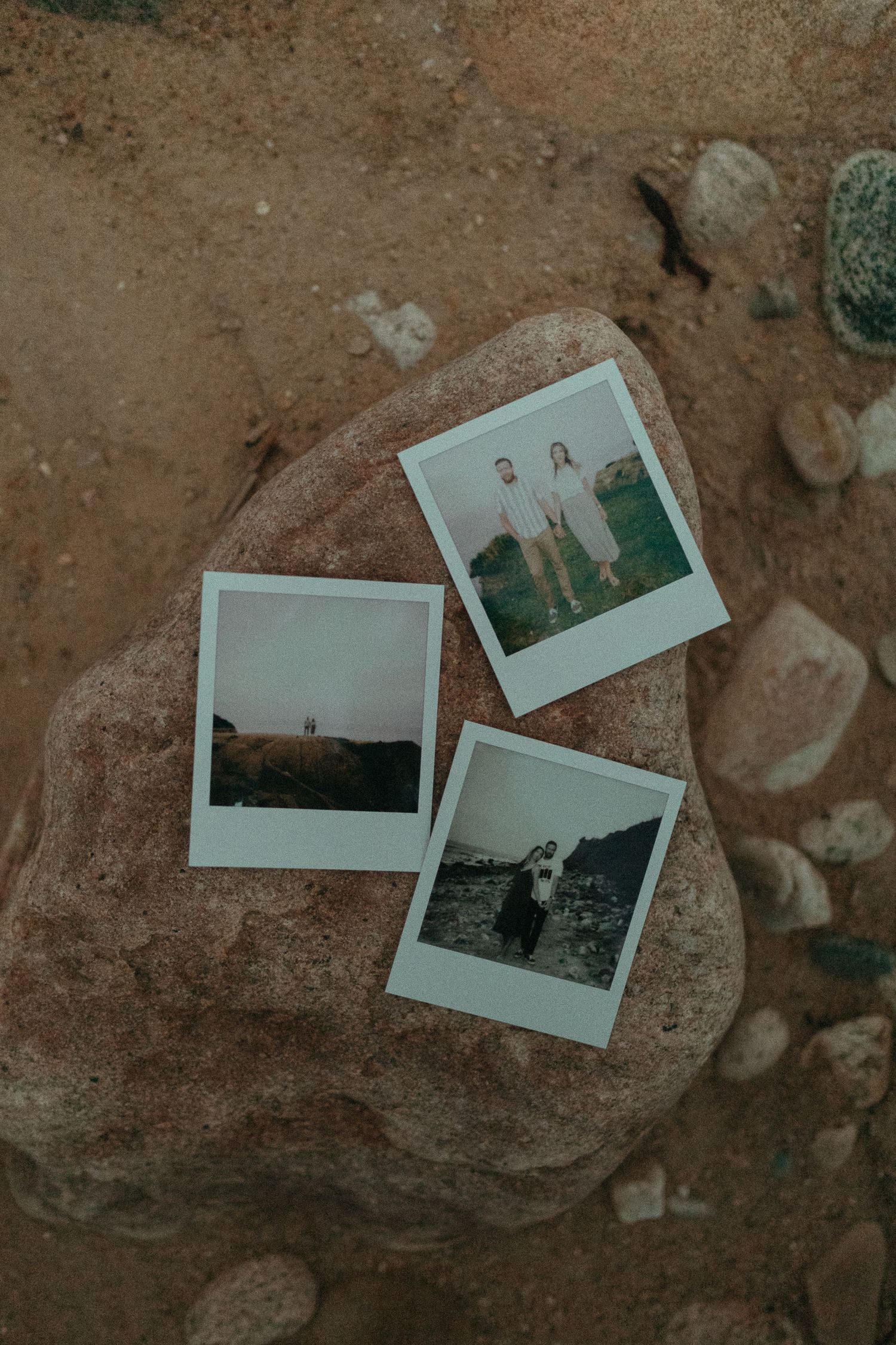 Three Polaroid photos of a couple arranged on a rocky beach surface with pebbles around them.