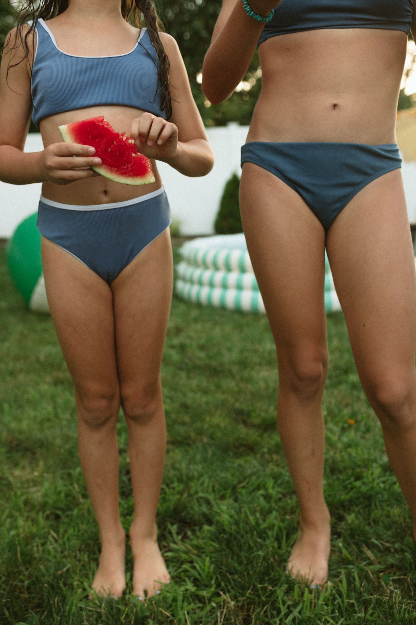 Two people in blue swimsuits holding watermelon slices in a backyard setting.