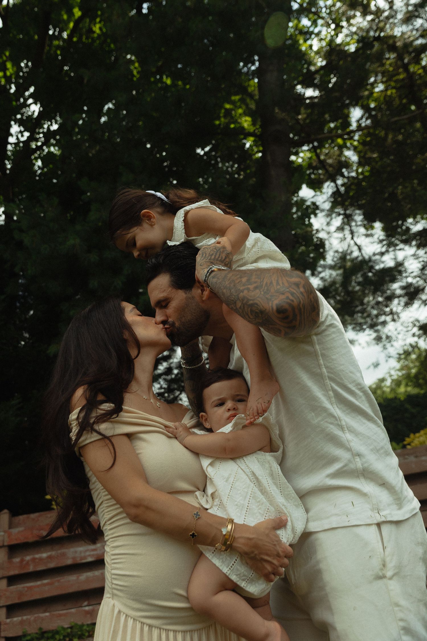 Tattooed couple kissing outdoors while holding two young children among green trees.