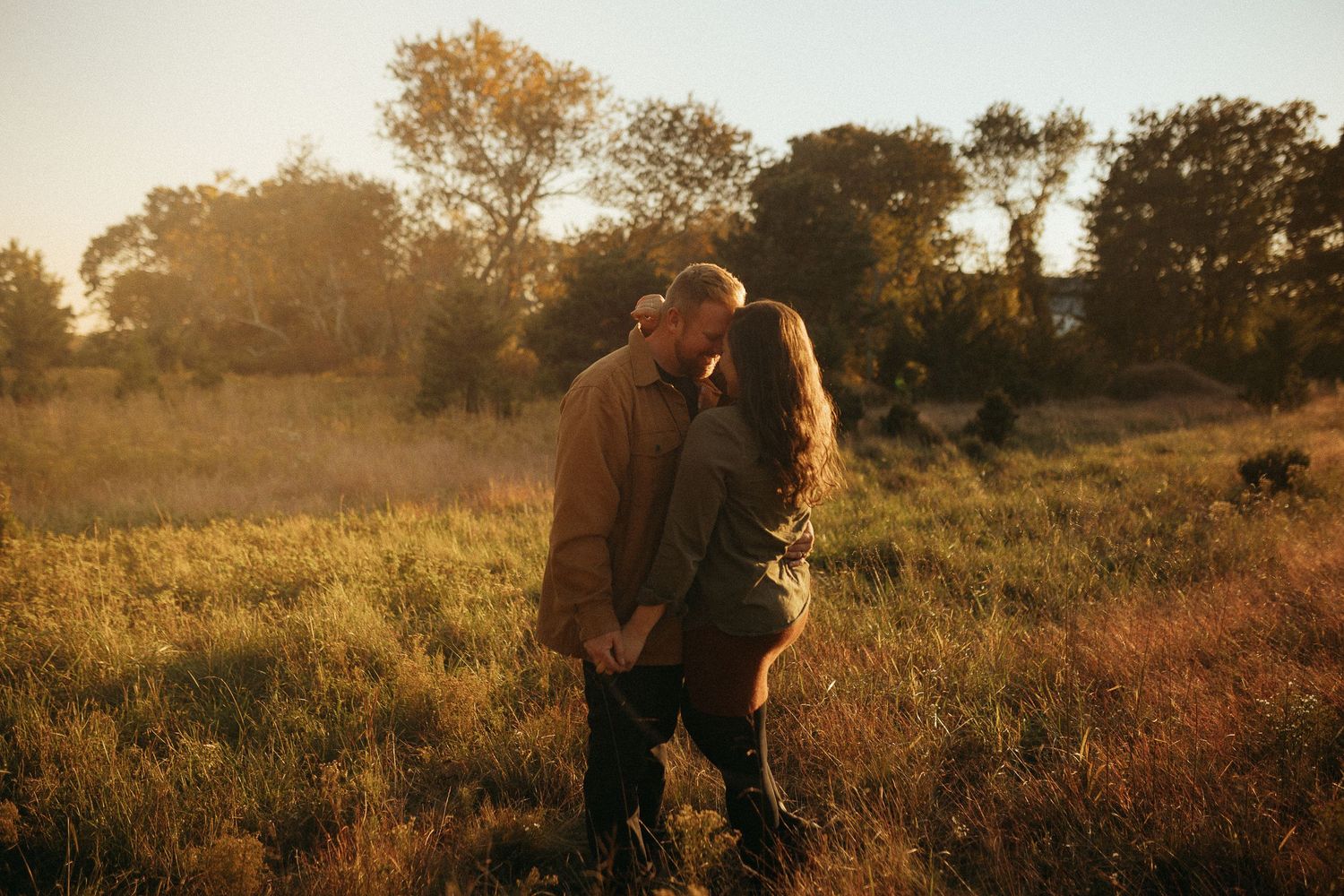 Couple sharing a romantic kiss in a sunlit golden meadow surrounded by lush green grass and trees at sunset.
