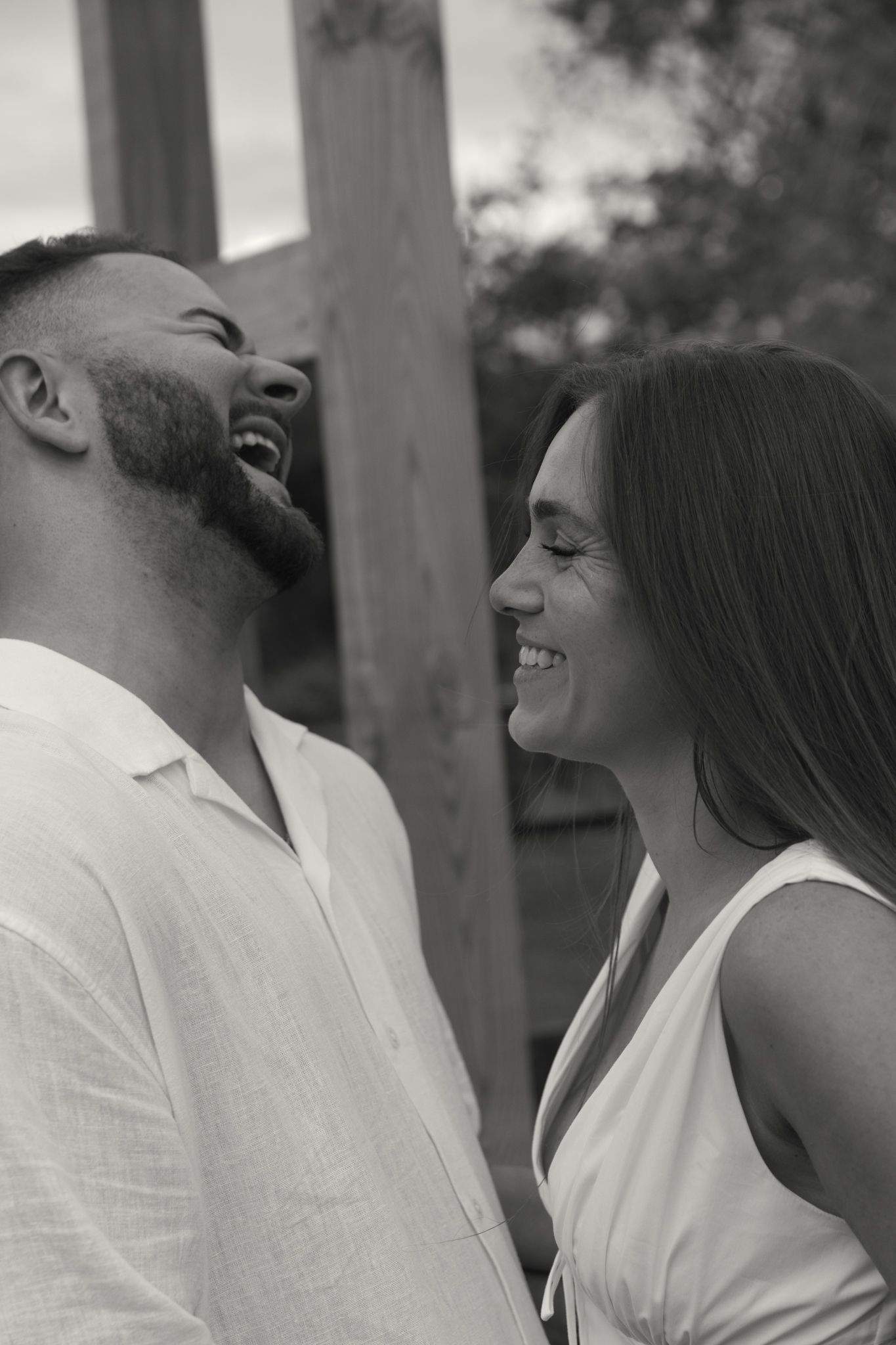 Black and white photo of a laughing couple in white outfits sharing a joyful moment outdoors.