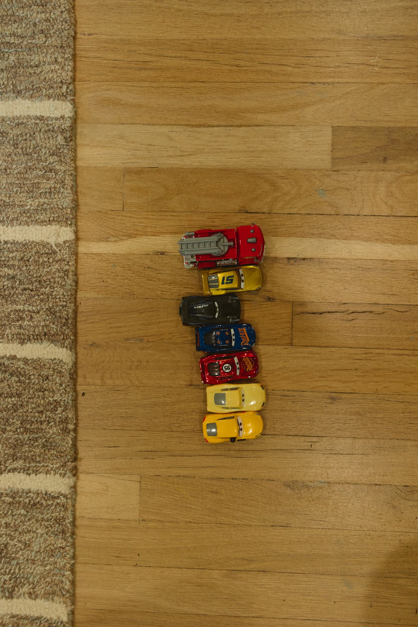 Colorful toy cars lined up on a wooden floor next to a beige rug.