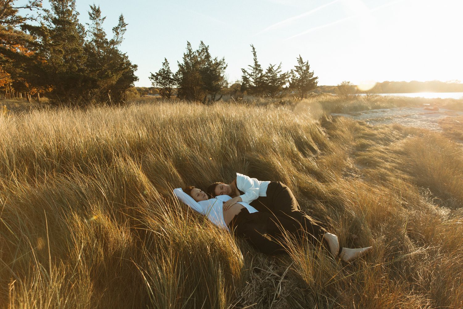 Couple lying together in golden tall grass field at sunset with trees and coastline in warm autumn light.