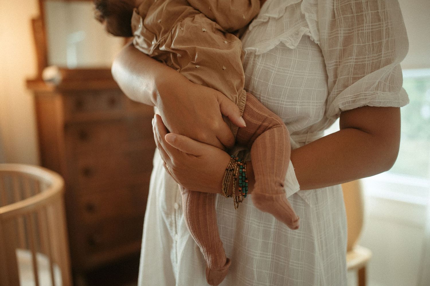 Mother in white lace dress tenderly holding a sleeping newborn baby in a warmly lit nursery.