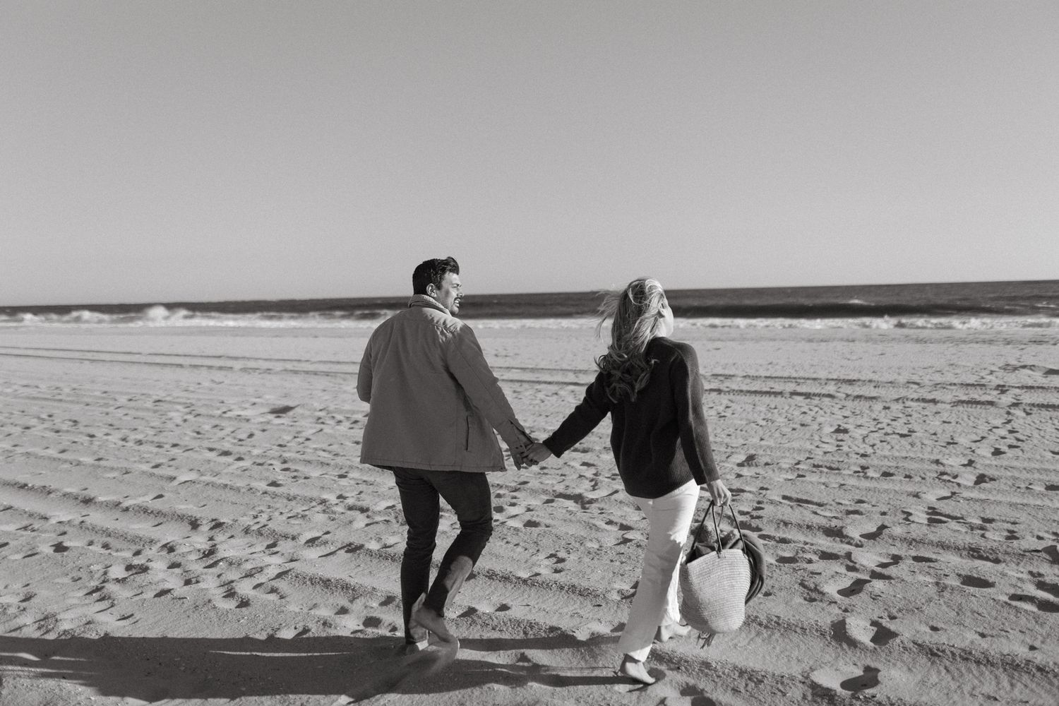 A couple runs hand-in-hand across a sandy beach in black and white.