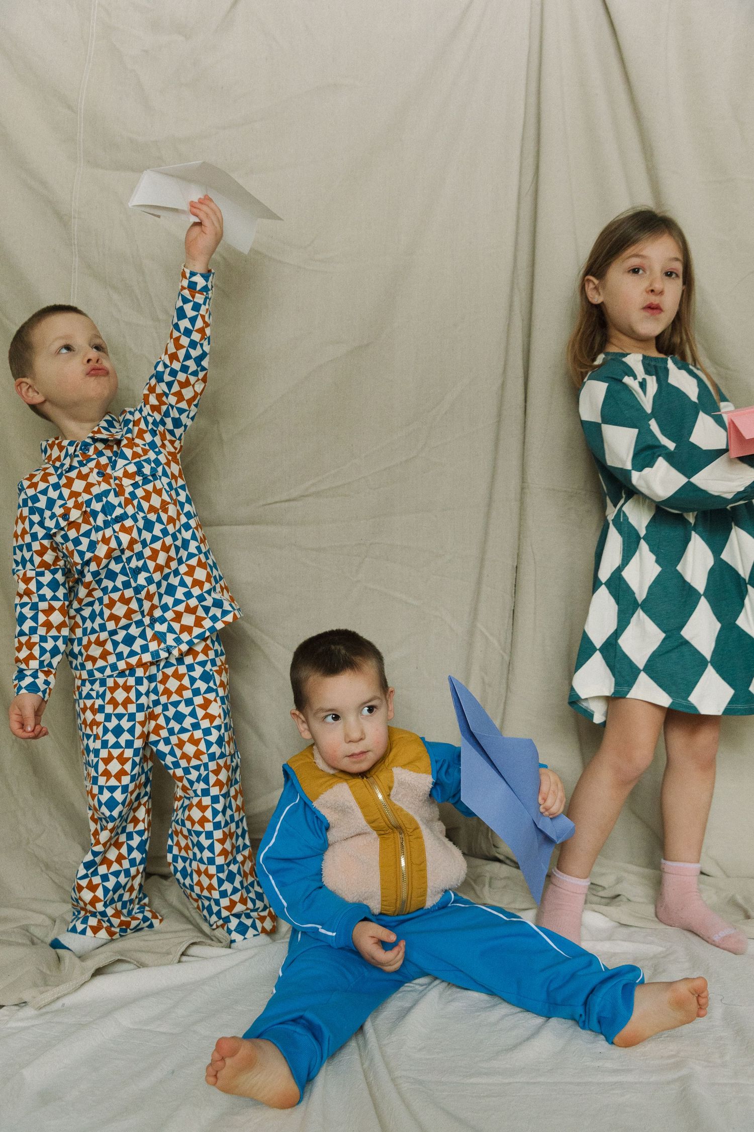 Three children in colorful patterned pajamas playing with paper airplanes against a neutral backdrop.