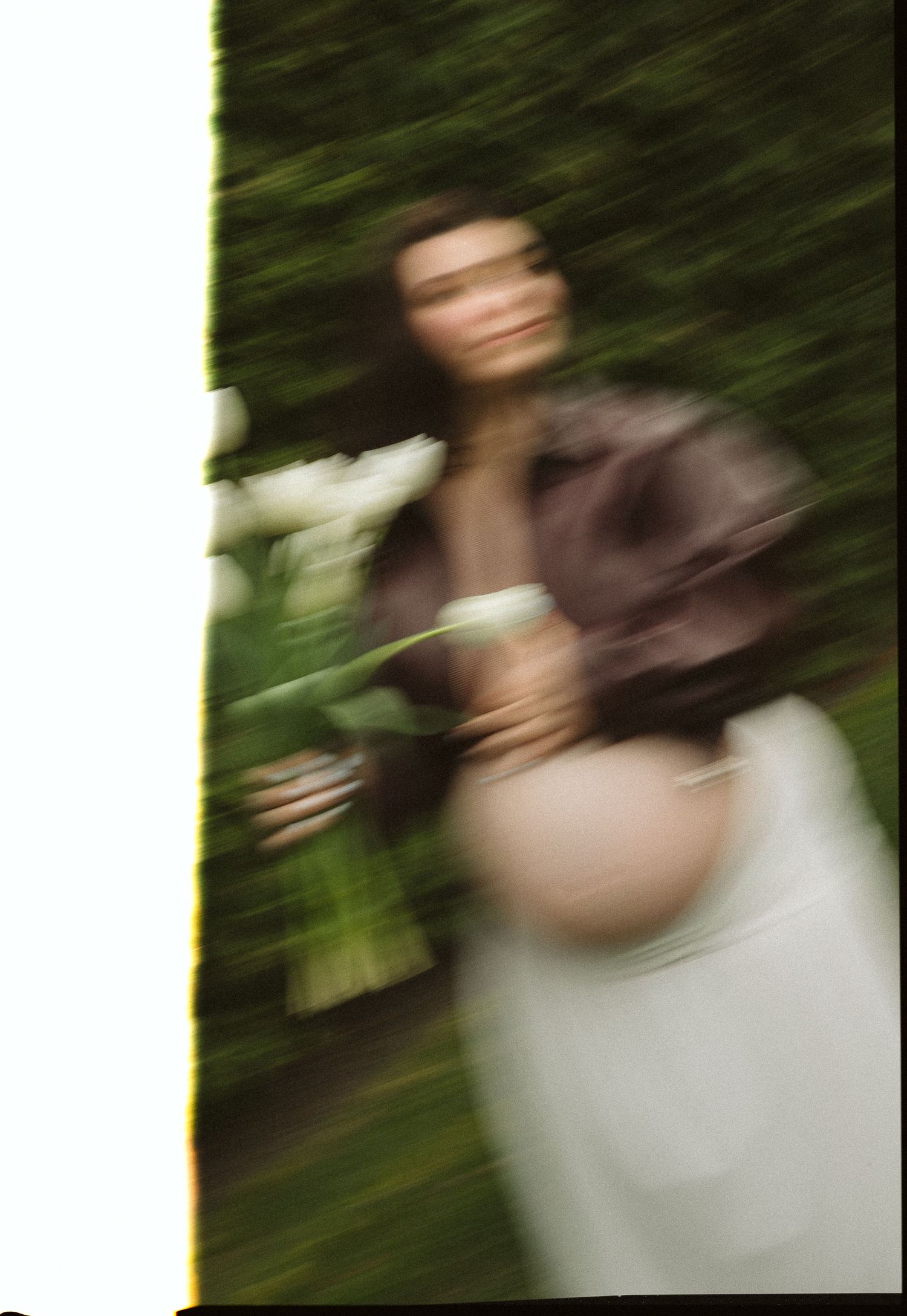 Blurred photo of a pregnant woman in a brown jacket holding white flowers near a green hedge.