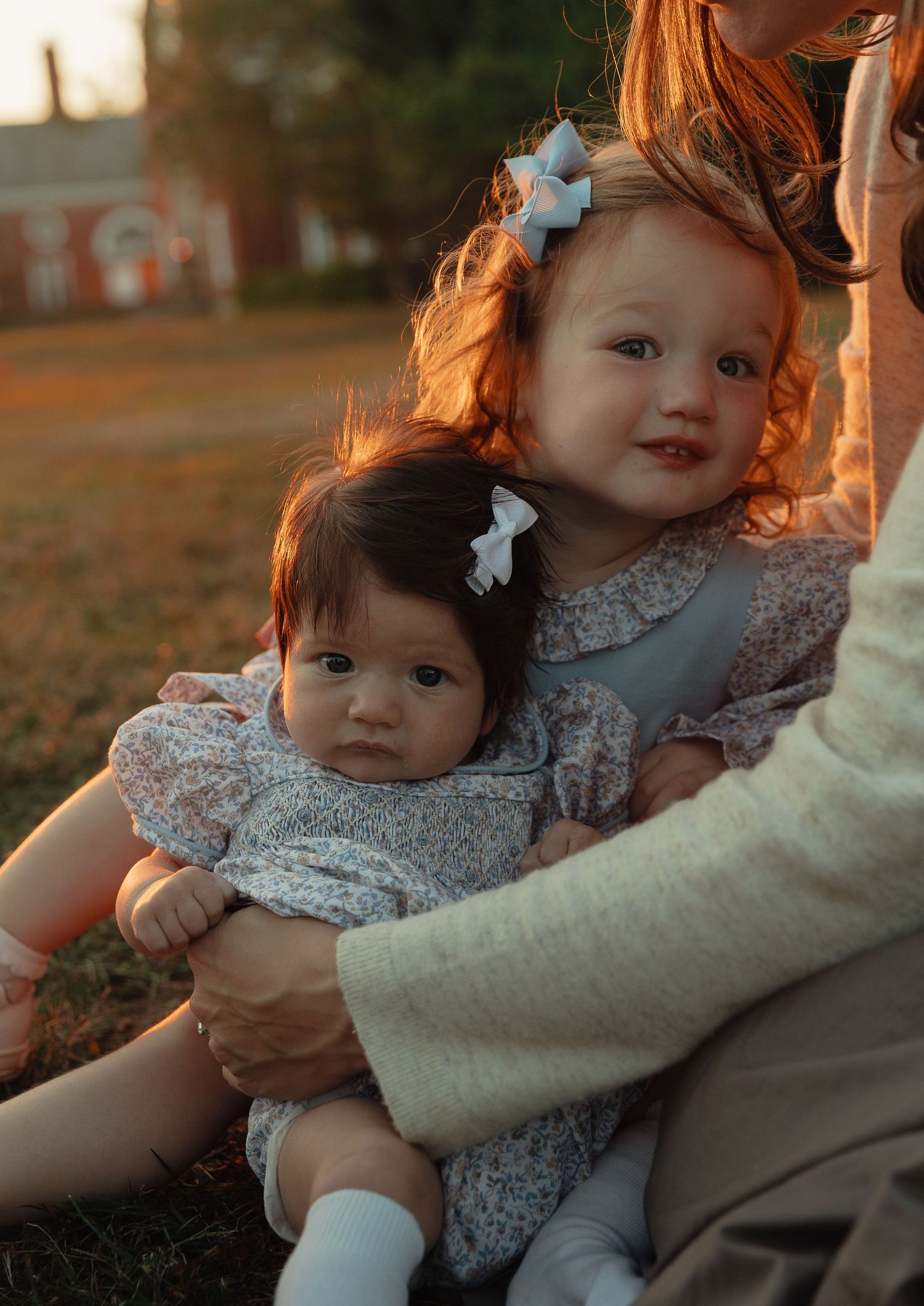 A toddler girl with a white bow holds a baby sibling while sitting outdoors at warm sunset light.