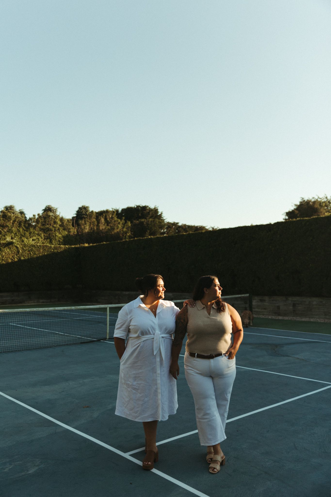 Two women walking together on a tennis court at golden hour, surrounded by lush green hedges.