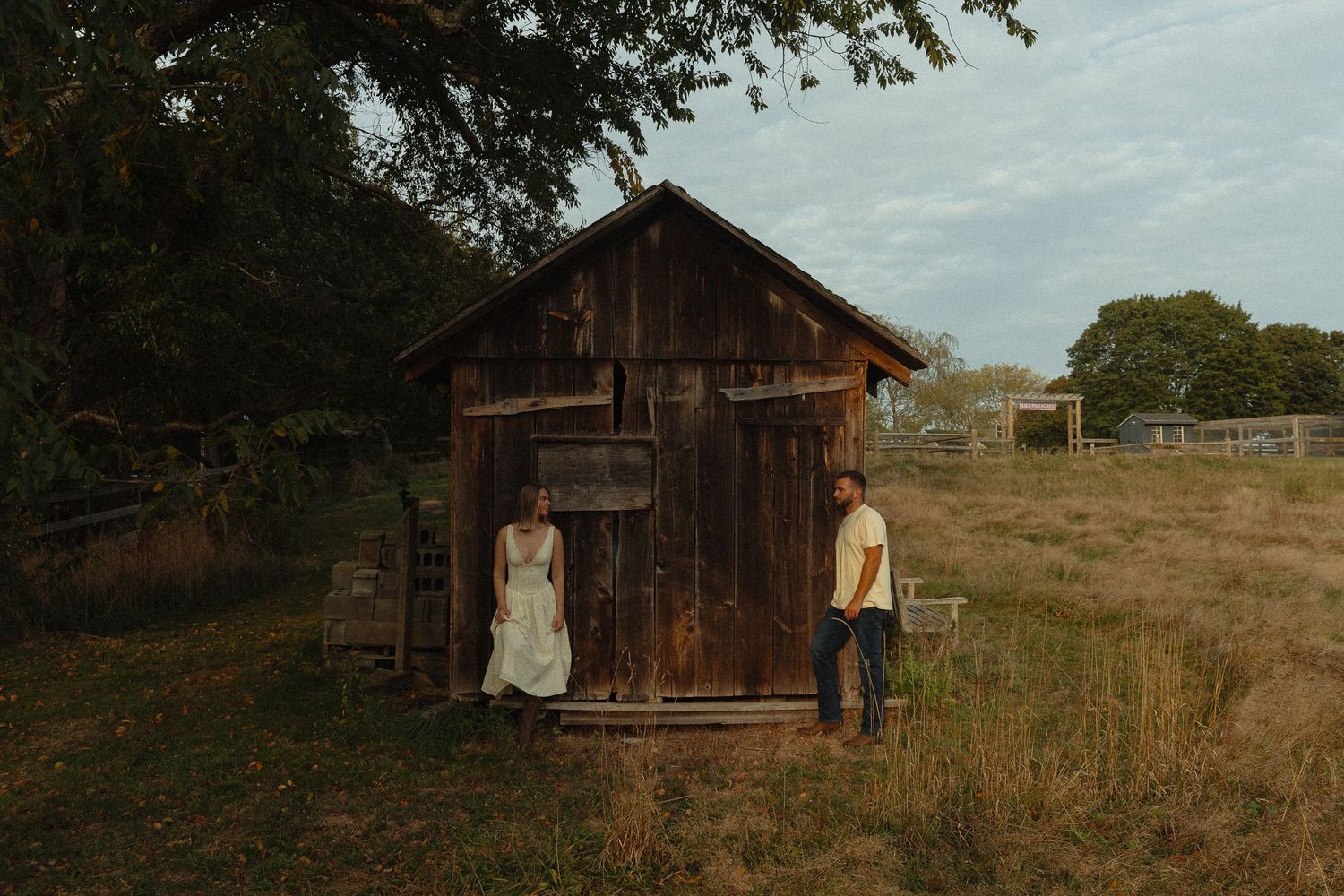 Couple standing apart by weathered wooden barn in golden afternoon light on rural farm.