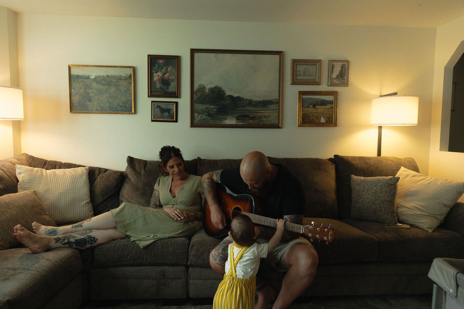 Family relaxing on couch as bald man plays acoustic guitar for woman and toddler in cozy living room.