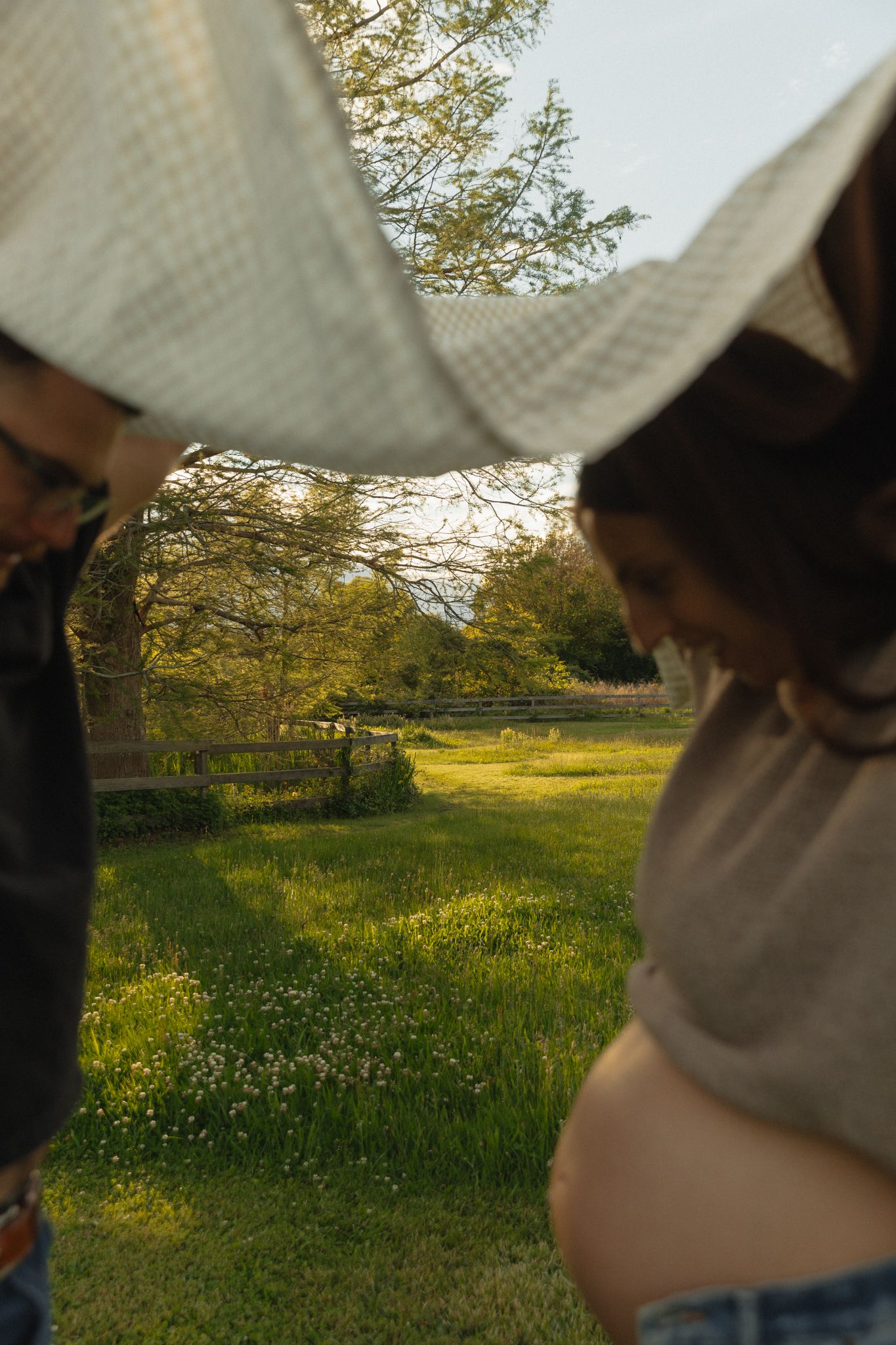 Pregnant woman standing outdoors in golden sunlight, with a blanket held above her revealing her baby bump in a green park.