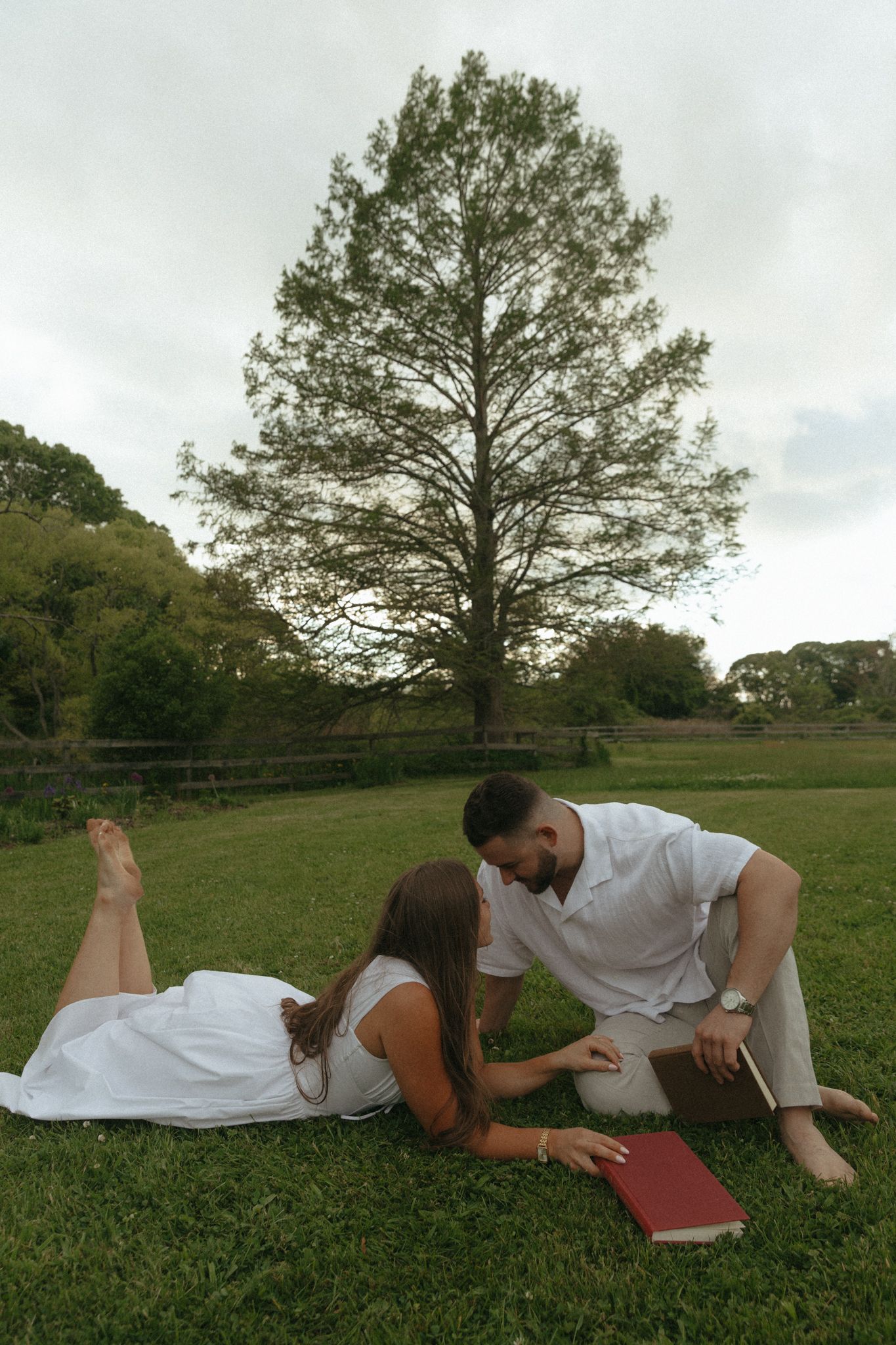Couple in white outfits sharing a romantic moment on green grass with a tall tree backdrop.