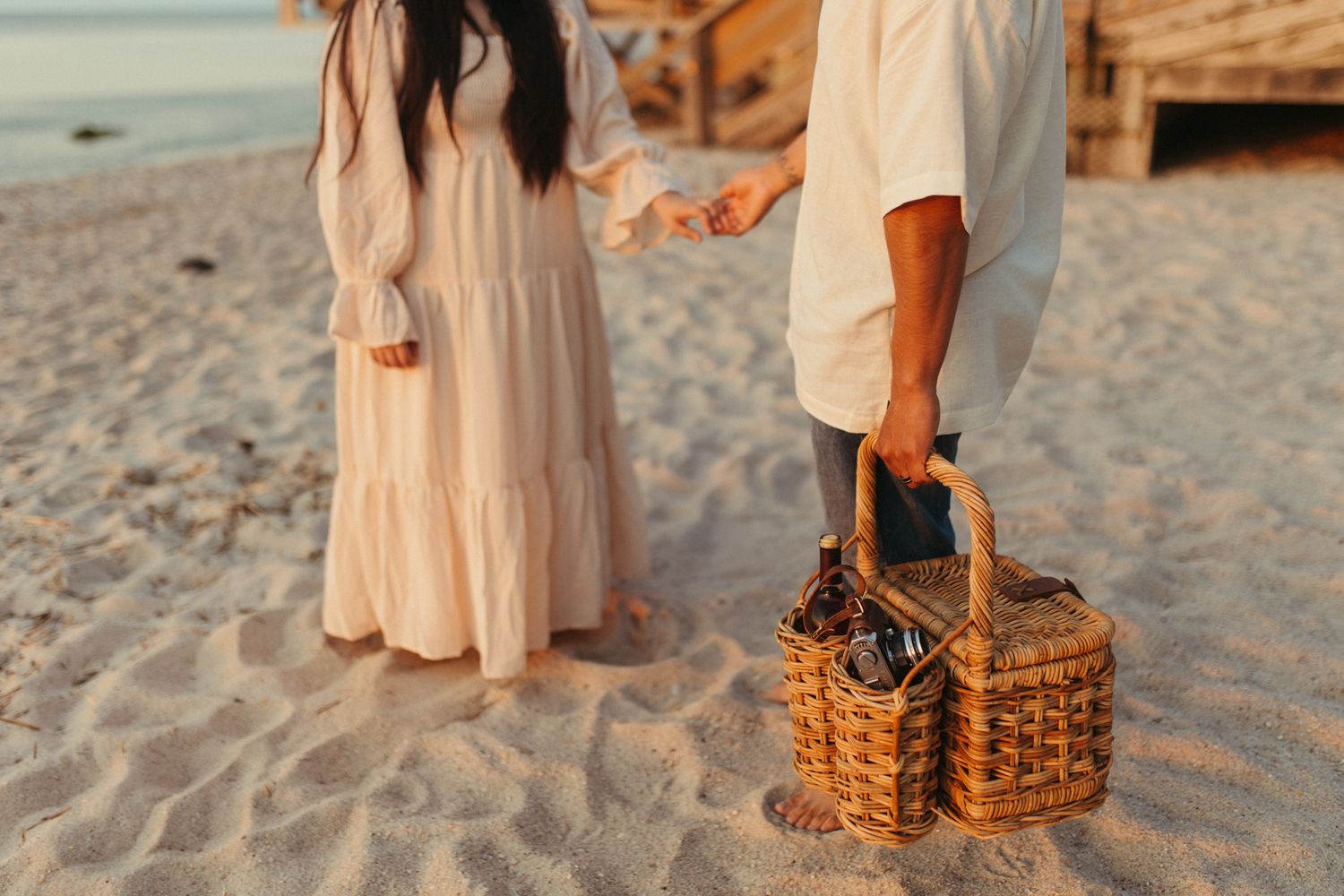 Couple on sandy beach holding wicker picnic basket at sunset, wearing white and cream boho outfits.