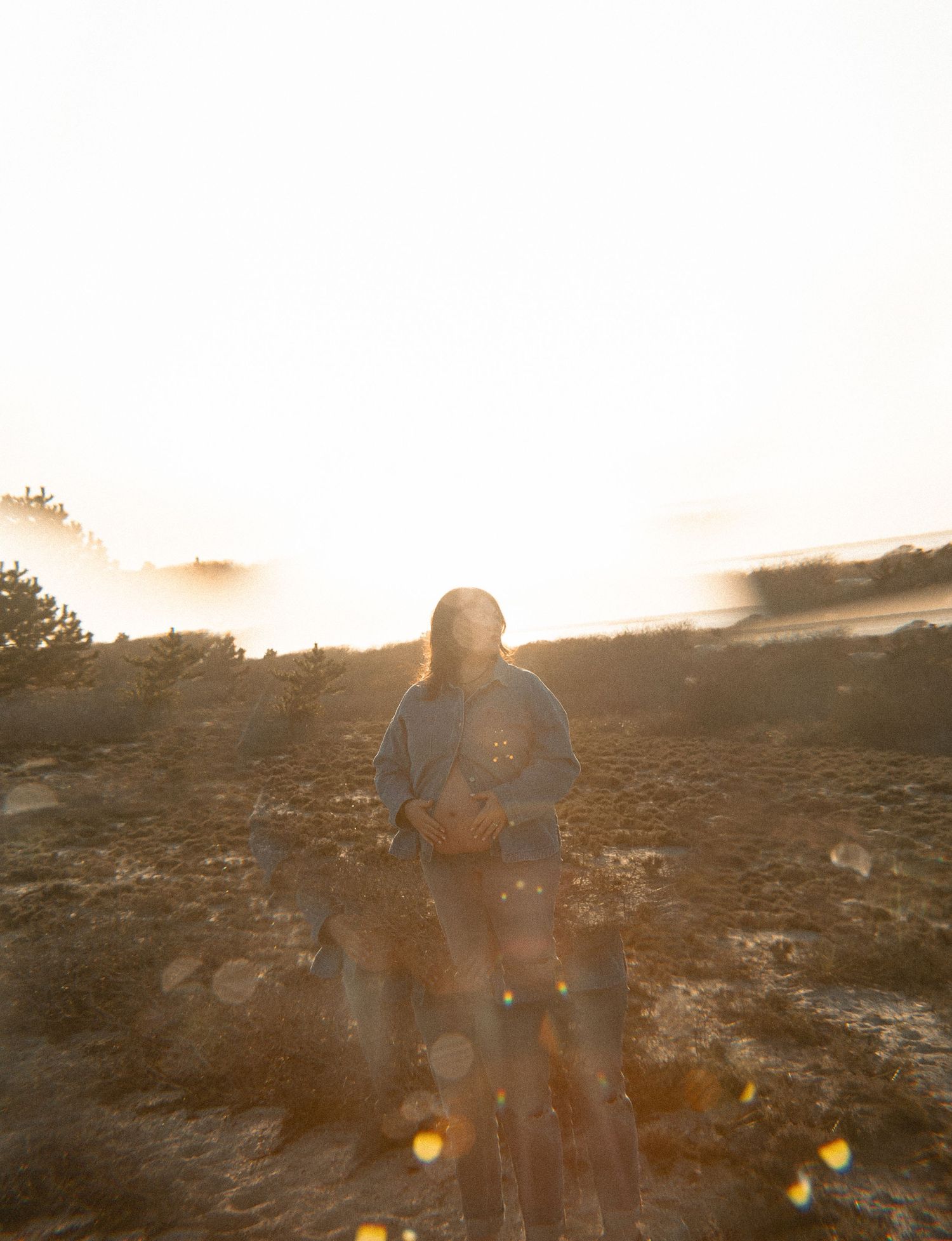 A woman stands in golden sunlight on a rocky landscape with misty horizon and lens flare.