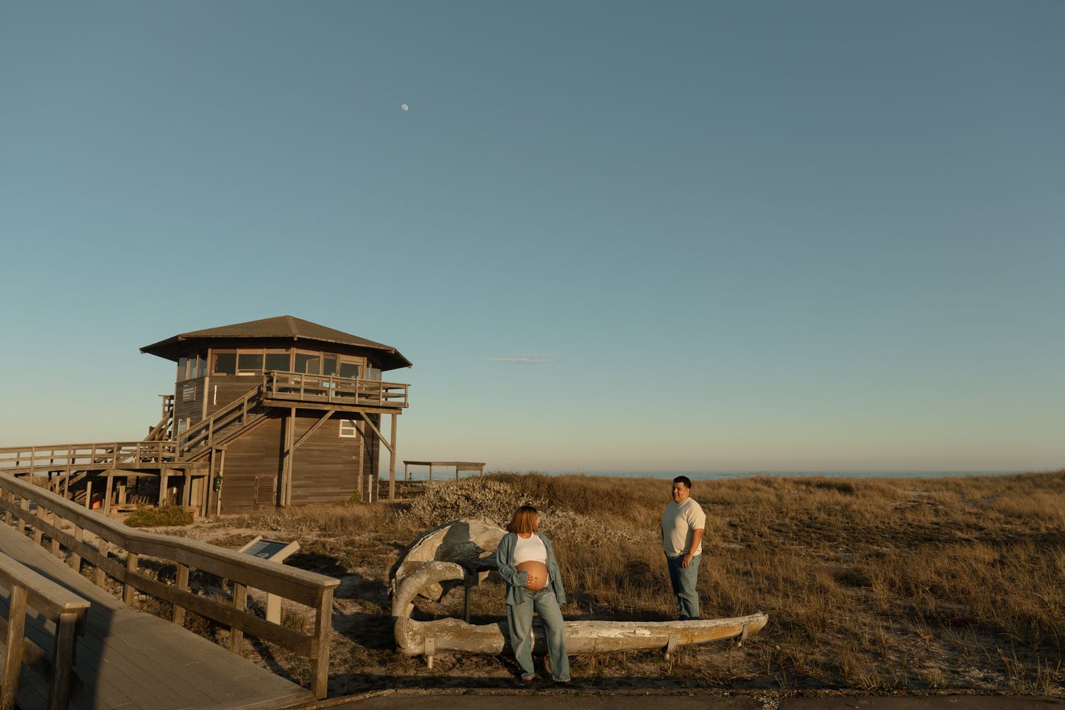 Couple standing near driftwood log beside coastal watchtower at golden hour sunset.