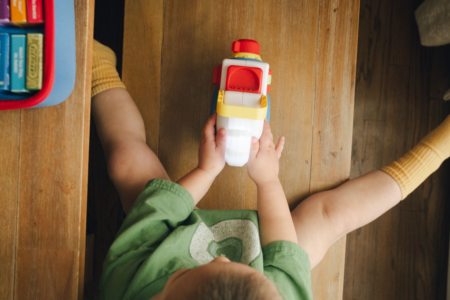Small hands holding a red and white toy bottle against a wooden cabinet background.