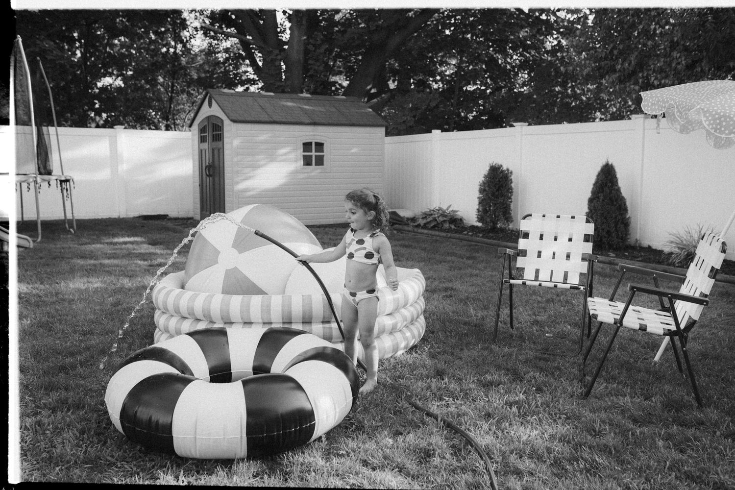 Black and white photo of a backyard with striped inflatable pool toys and white fence on a sunny day.