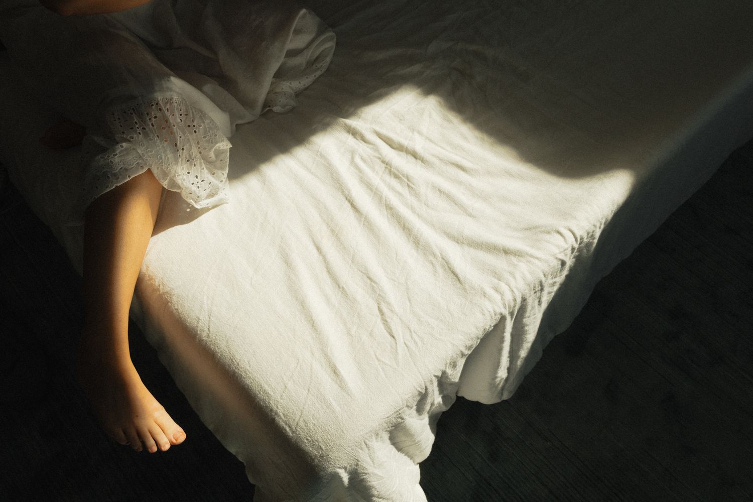 Soft morning light casting shadows over woman in white lace dress sitting on edge of bed.