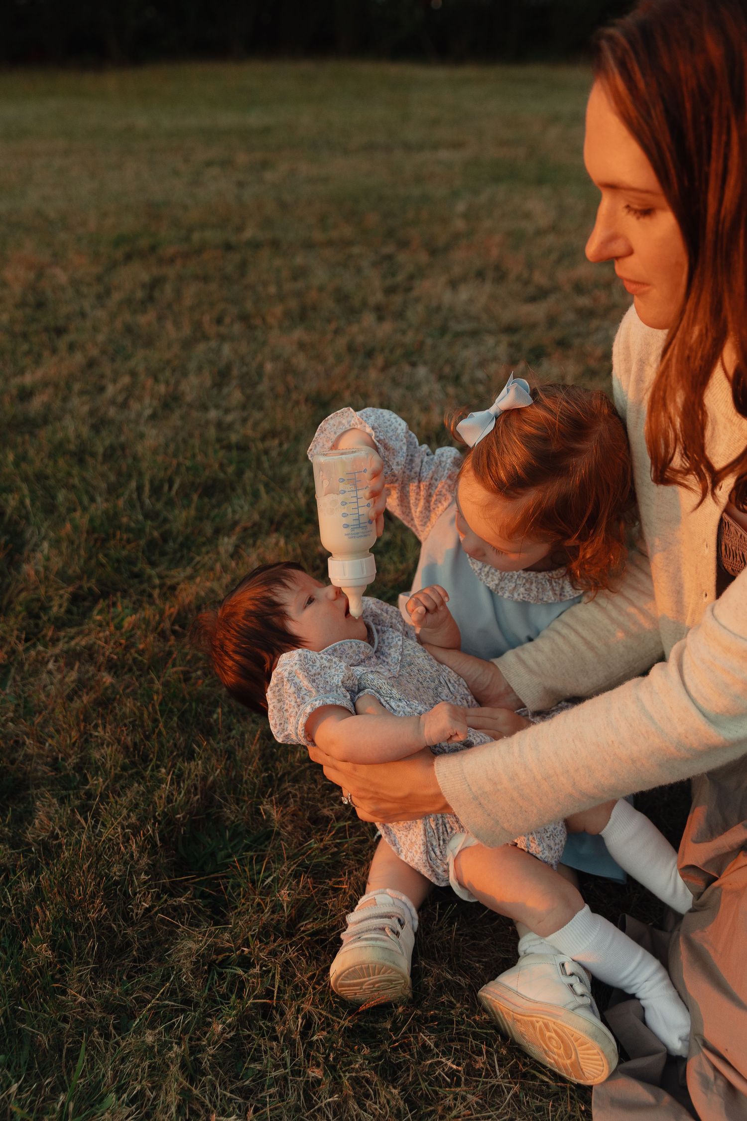 A mother sits on grass at sunset, bottle-feeding her baby while a toddler girl in a blue dress helps hold the bottle.