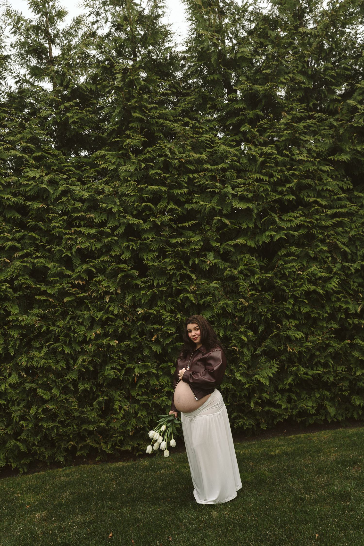Pregnant woman in white skirt and brown jacket holding white tulips, standing in front of lush green hedge outdoors.