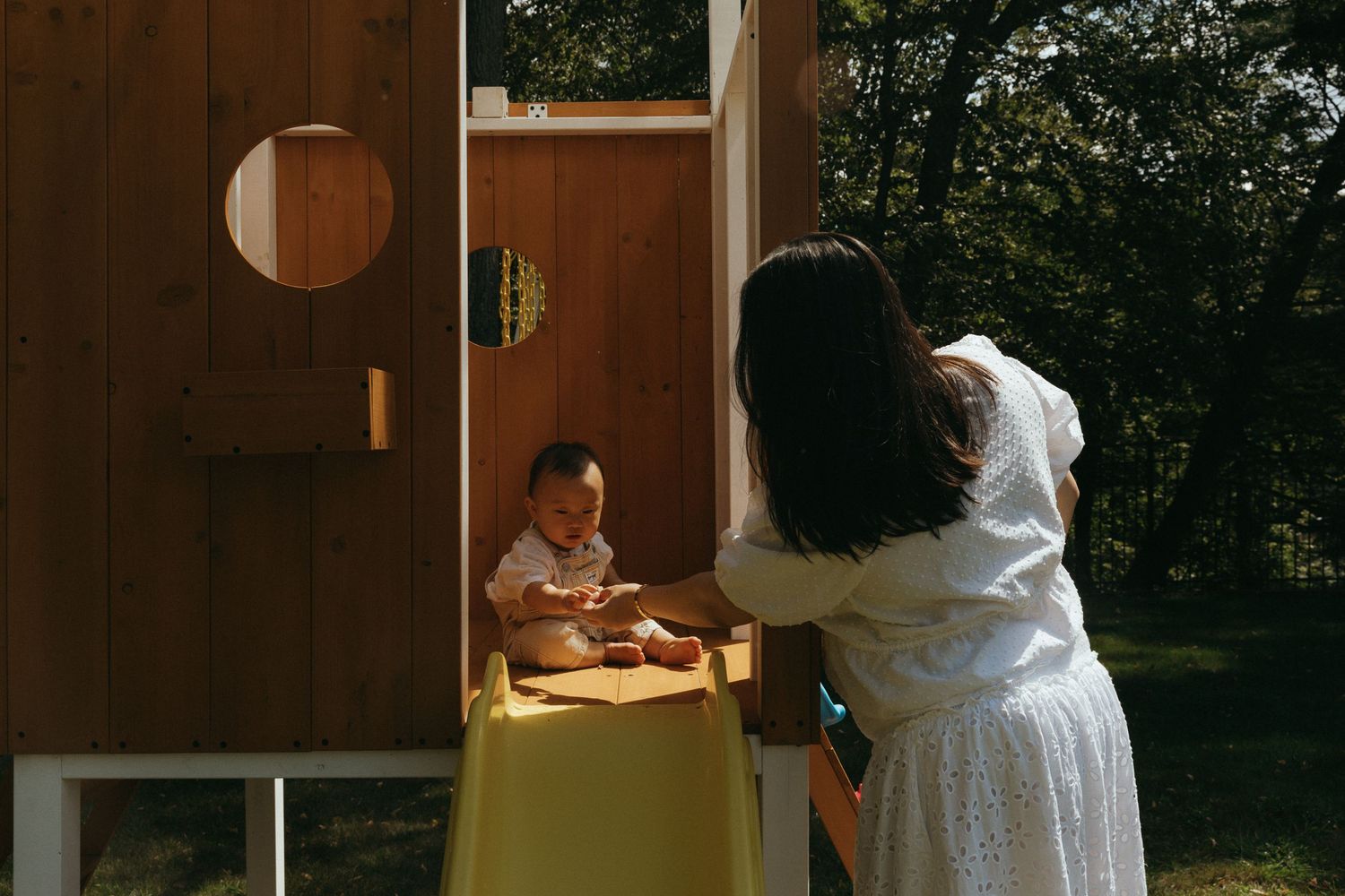 A mother in a white dress interacts with her baby sitting in a wooden playhouse with a yellow slide outdoors.