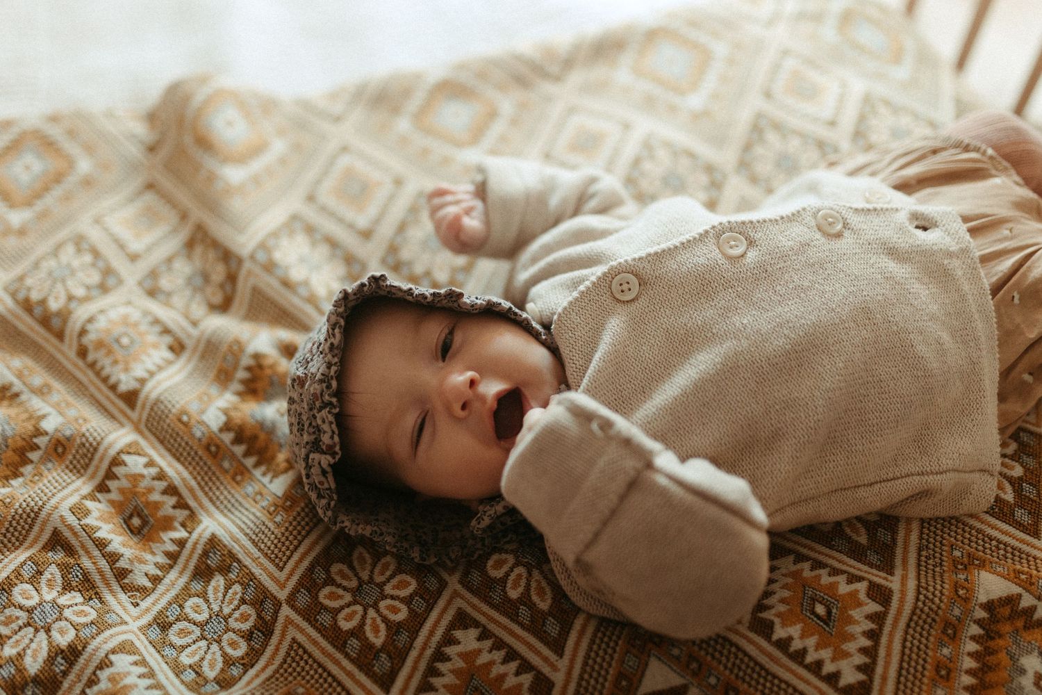 Newborn baby in beige knit outfit and floral bonnet lying on a patterned brown blanket.