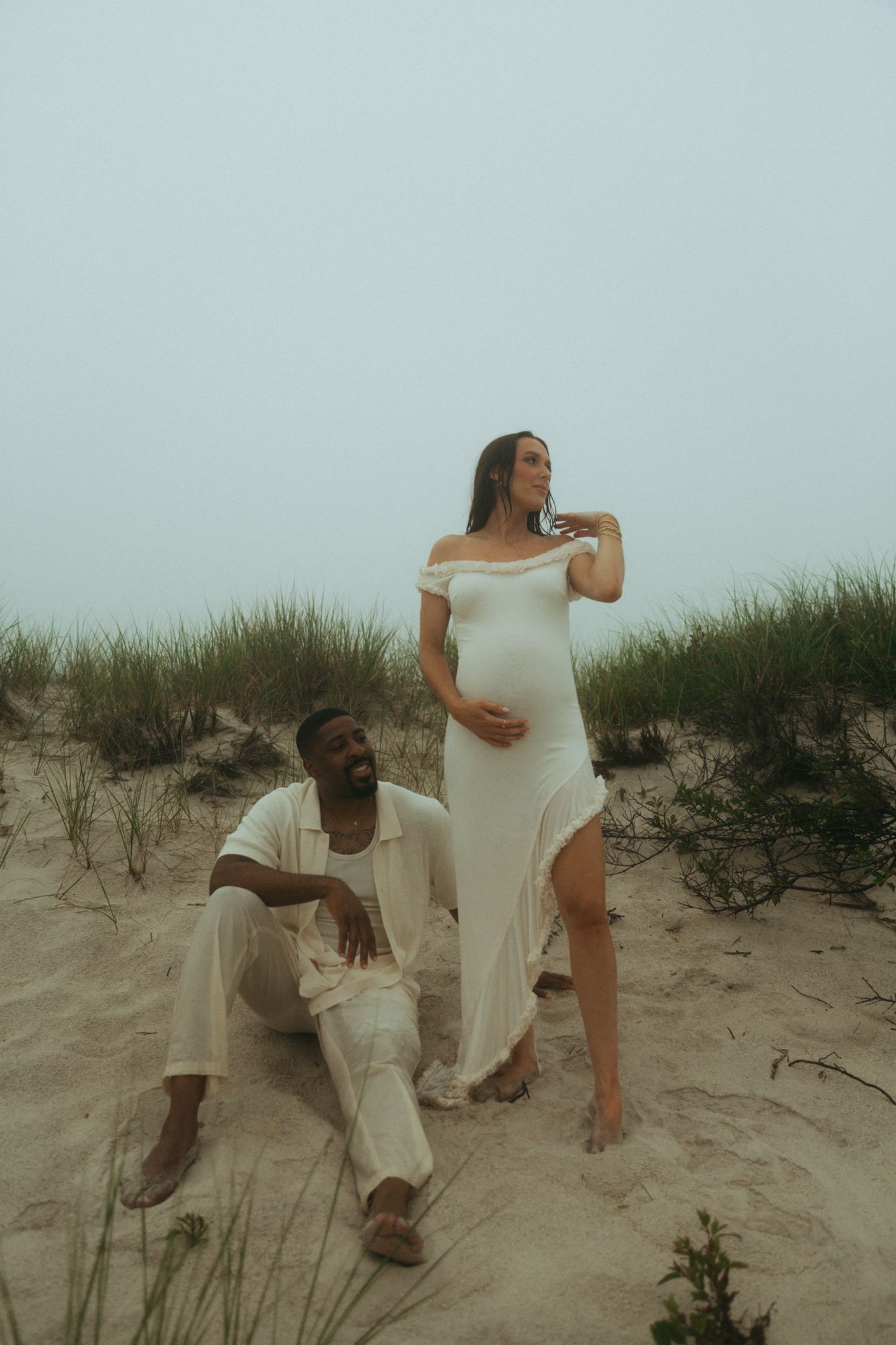 Couple in white outfits playfully interacting on sandy beach dunes.