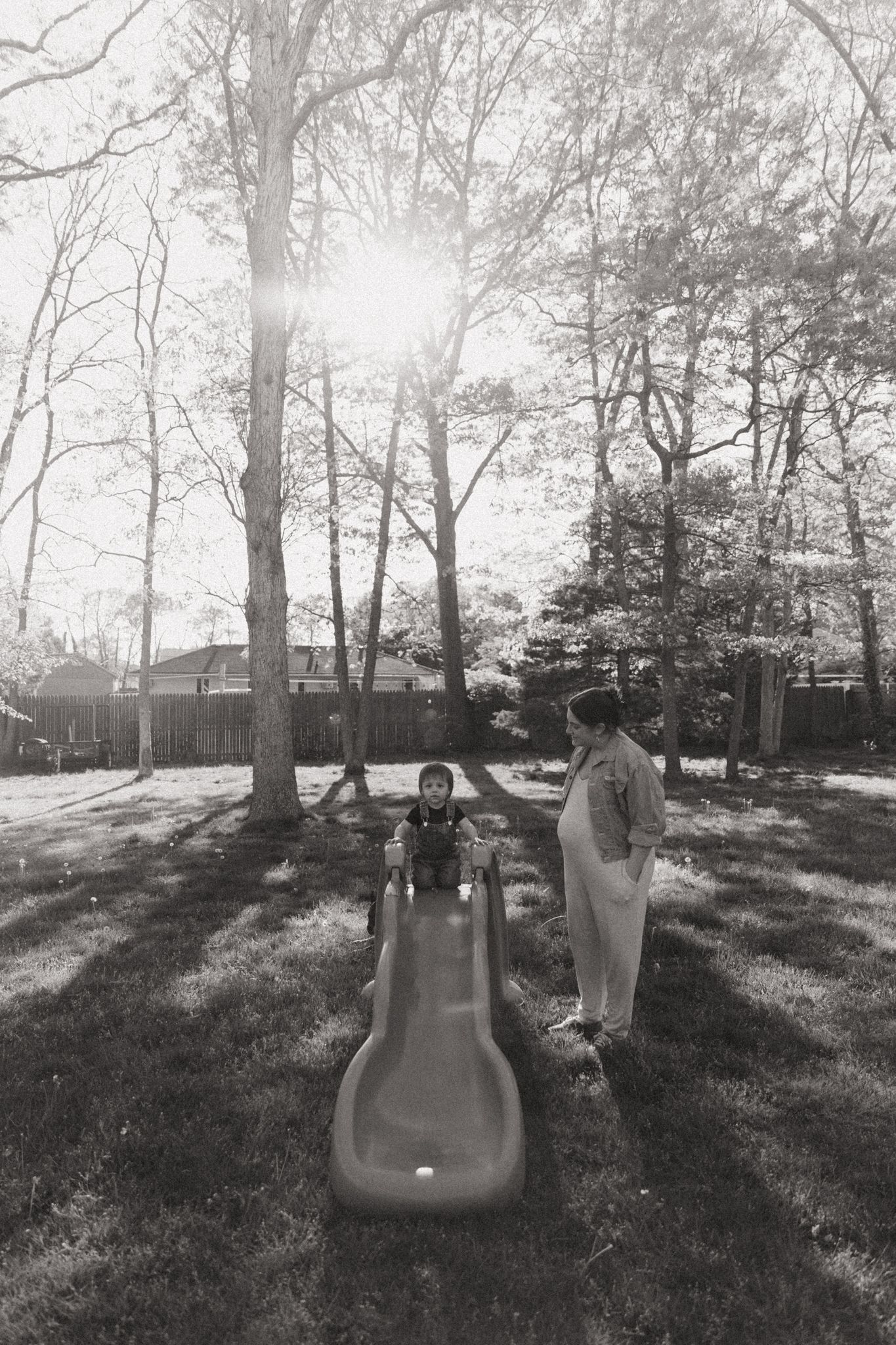 A black and white photo of a backyard playground with a slide and trees casting long shadows.