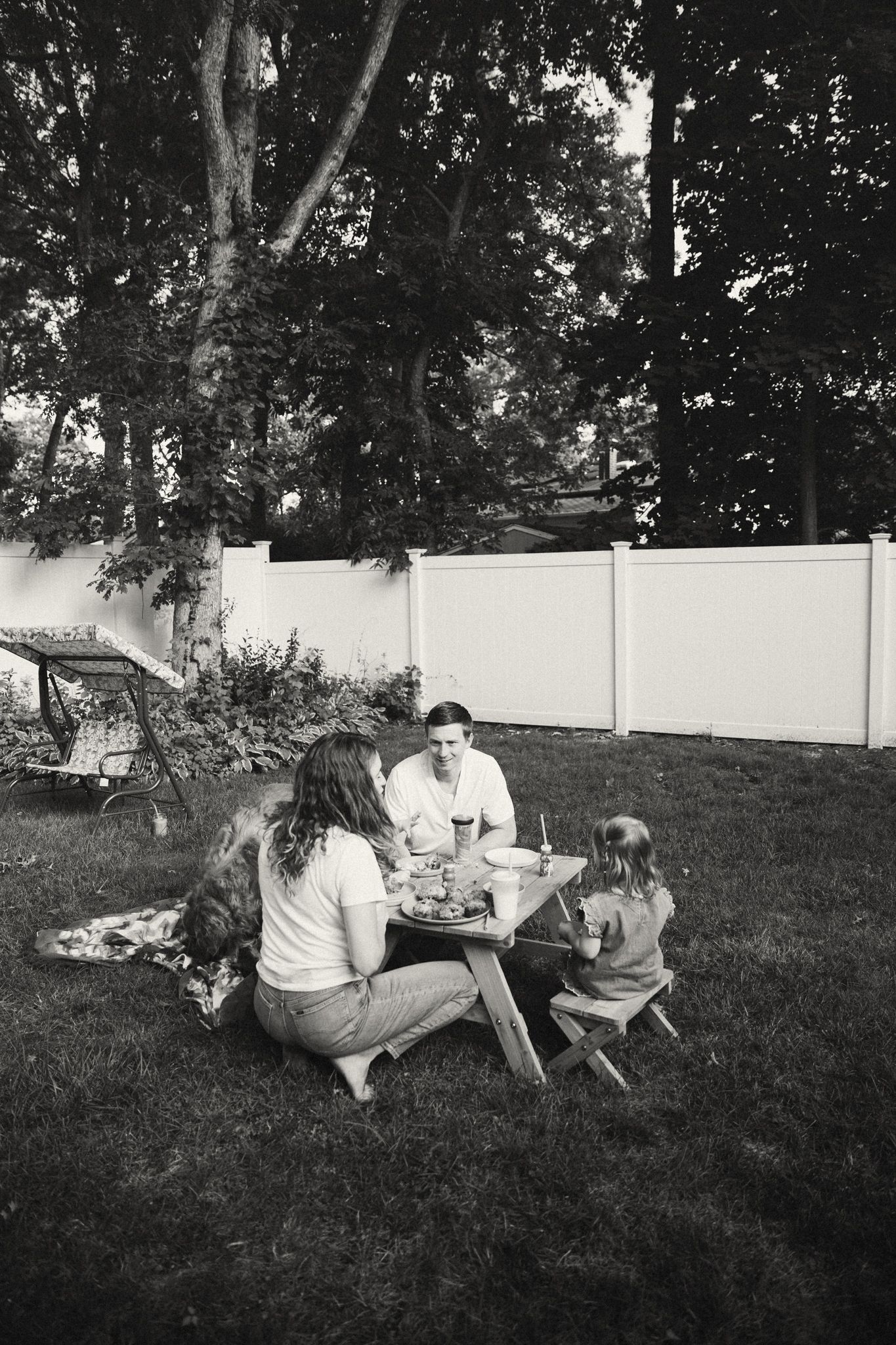 Black and white photo of a family enjoying a meal at a picnic table in their backyard.