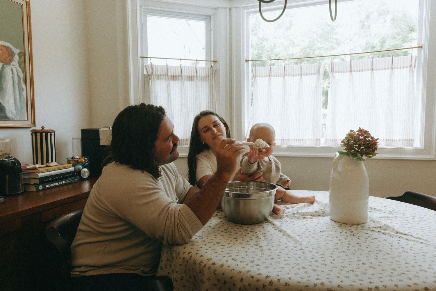 Two parents feeding their baby at a dining table near bright windows in a cozy home setting.