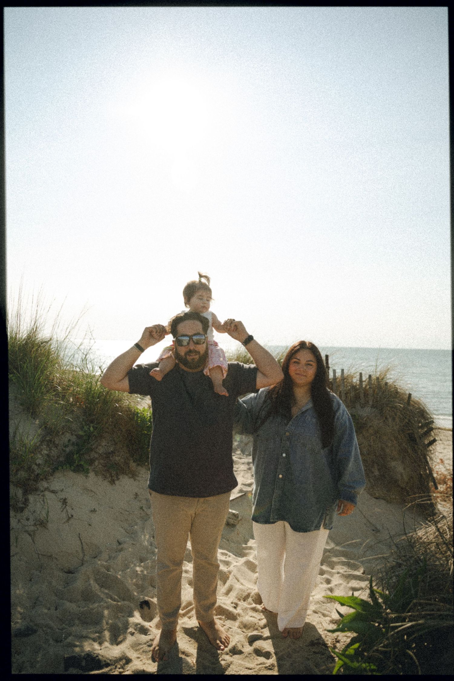Family of three with toddler on fathers shoulders posing on a sunny sandy beach.