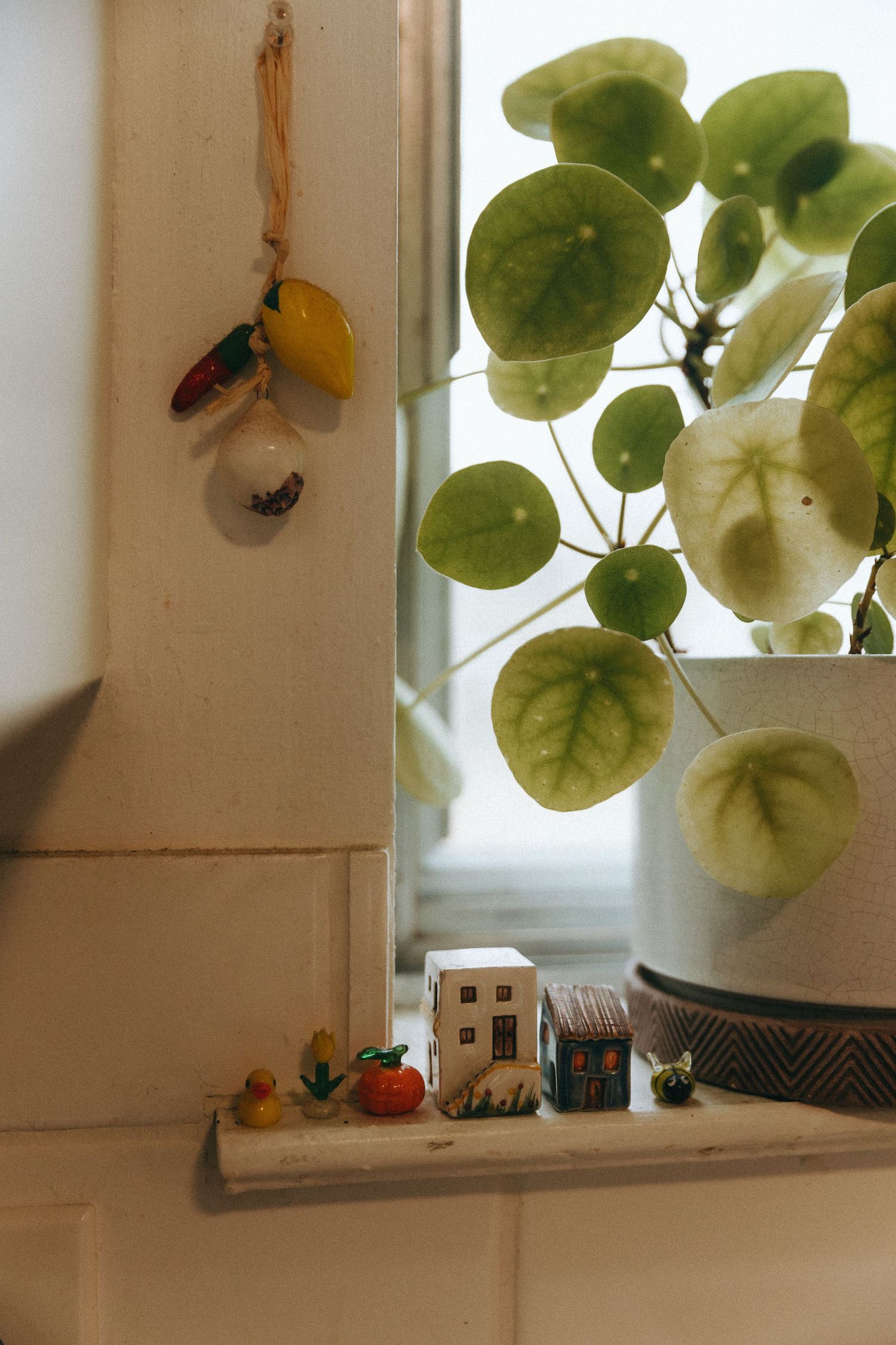 Pilea plant on sunny windowsill with miniature ceramic houses and tiny figurines on ledge.