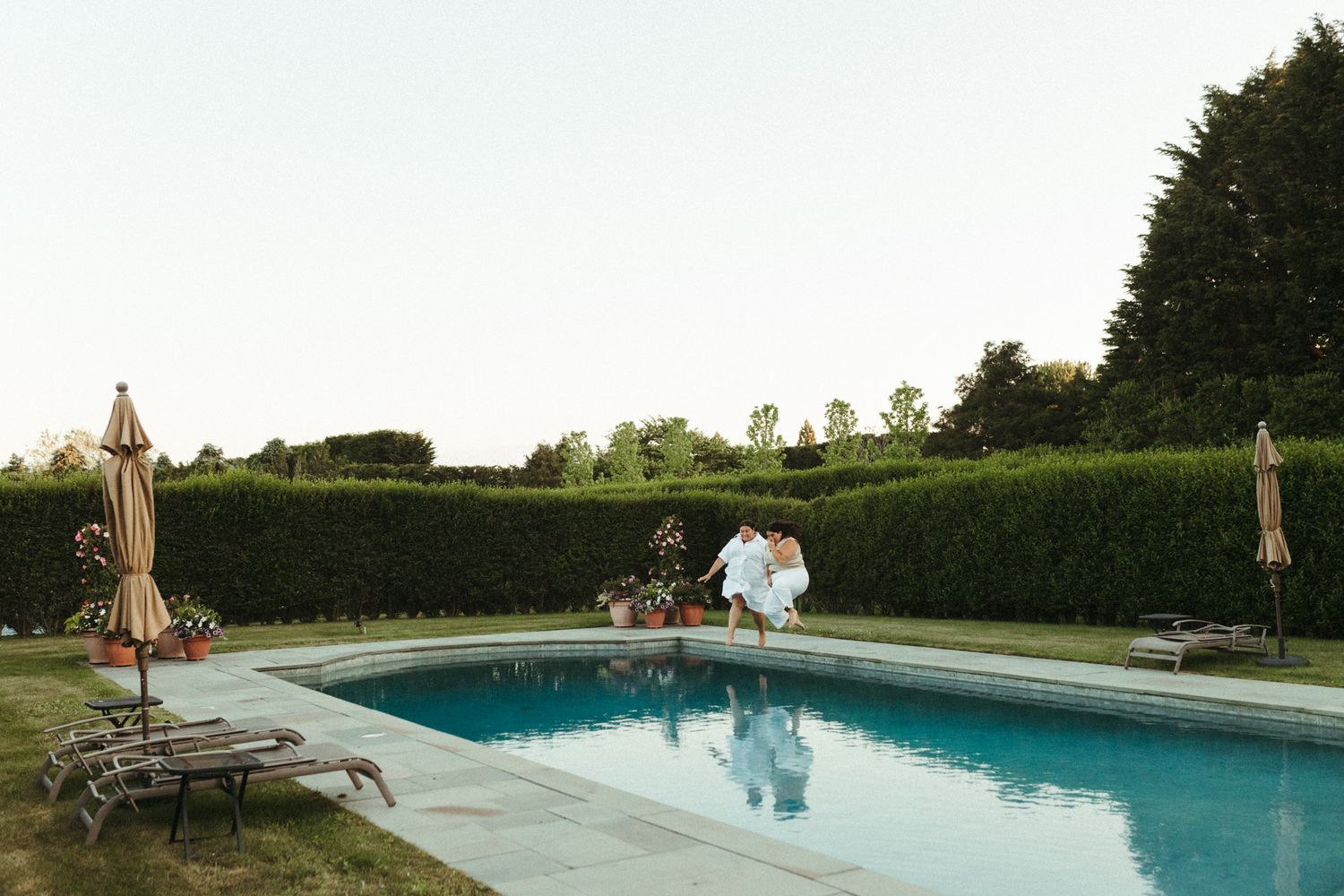 Couple embracing poolside at elegant estate with manicured hedges, lounge chairs, and patio umbrellas at dusk.