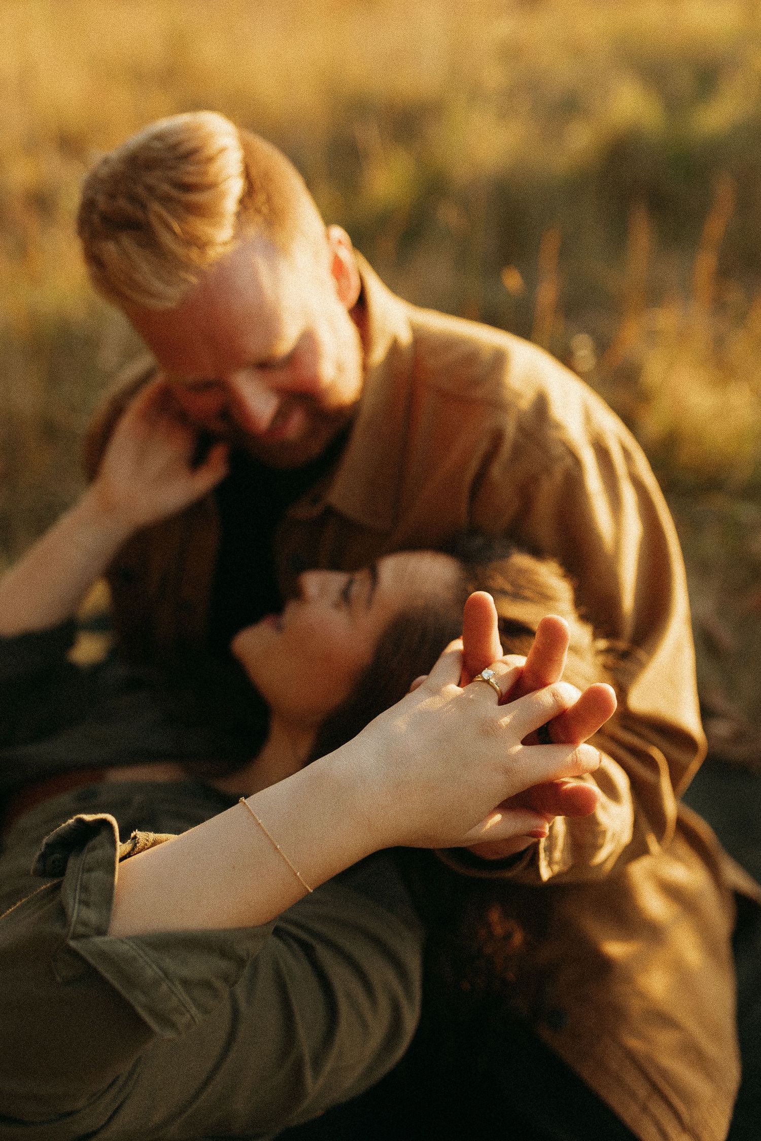 A couple shares a tender moment outdoors at golden hour, holding hands and smiling at each other in a sunlit field.
