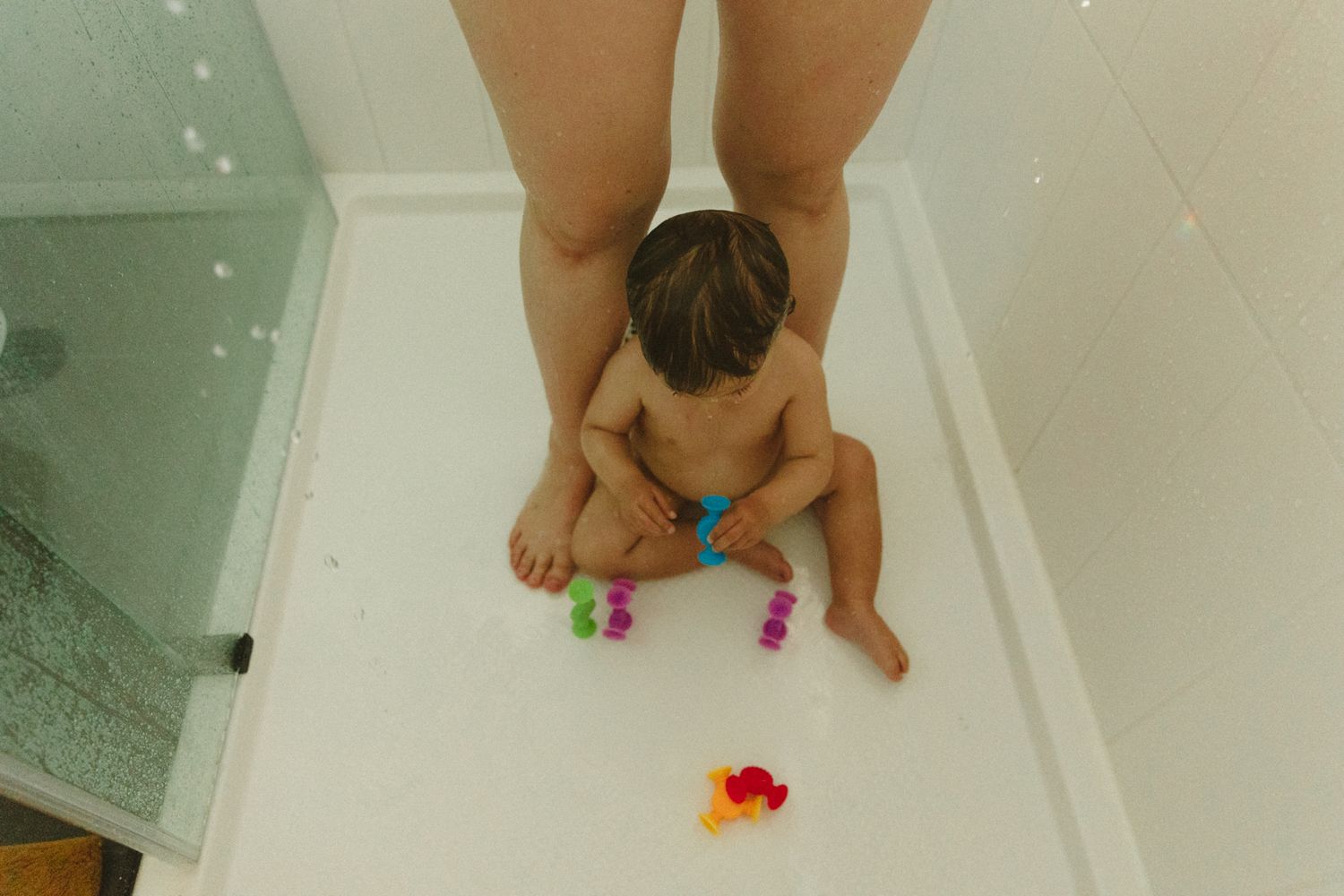 Baby sitting on shower floor with colorful toys while adult stands nearby during bath time.