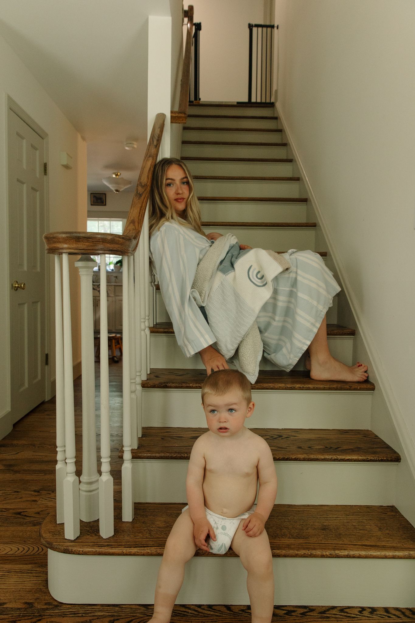 Smiling mother in striped robe sitting on wooden staircase with baby in diaper on bottom step.