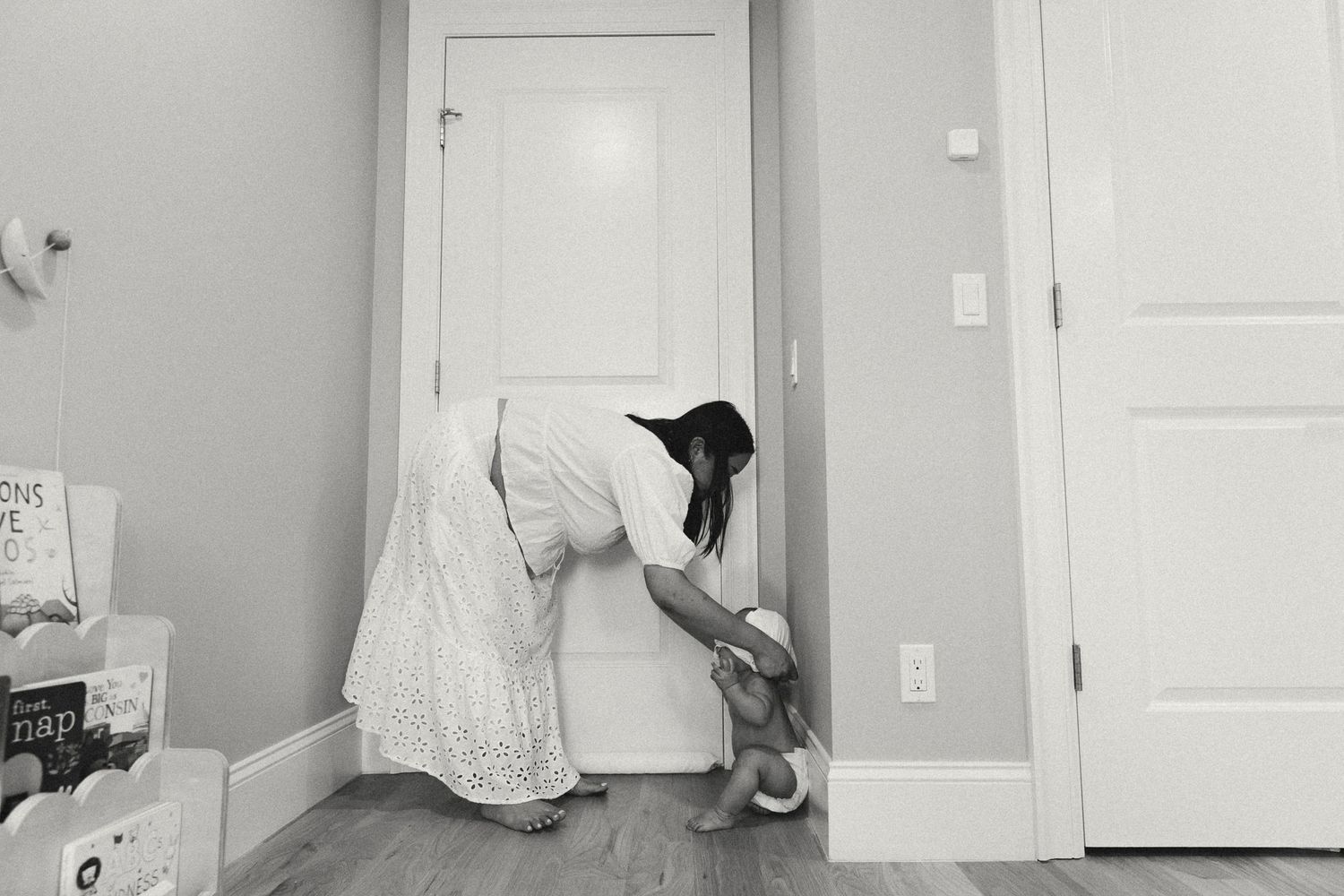 Black and white photo of mother in white dress bending down to tend to baby near doorway.