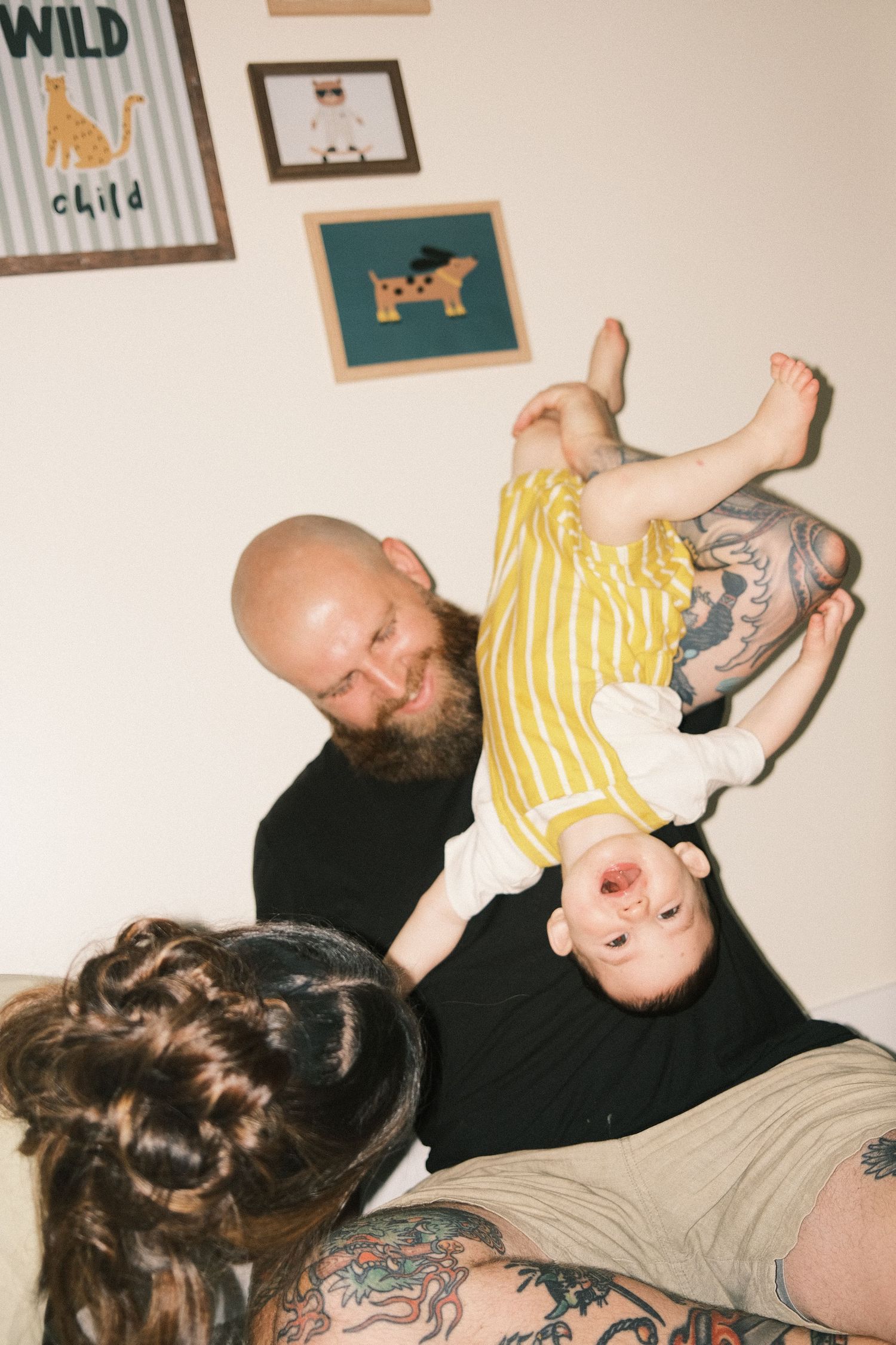 Tattooed parents playfully hold baby upside down in a cozy nursery with animal-themed wall art.