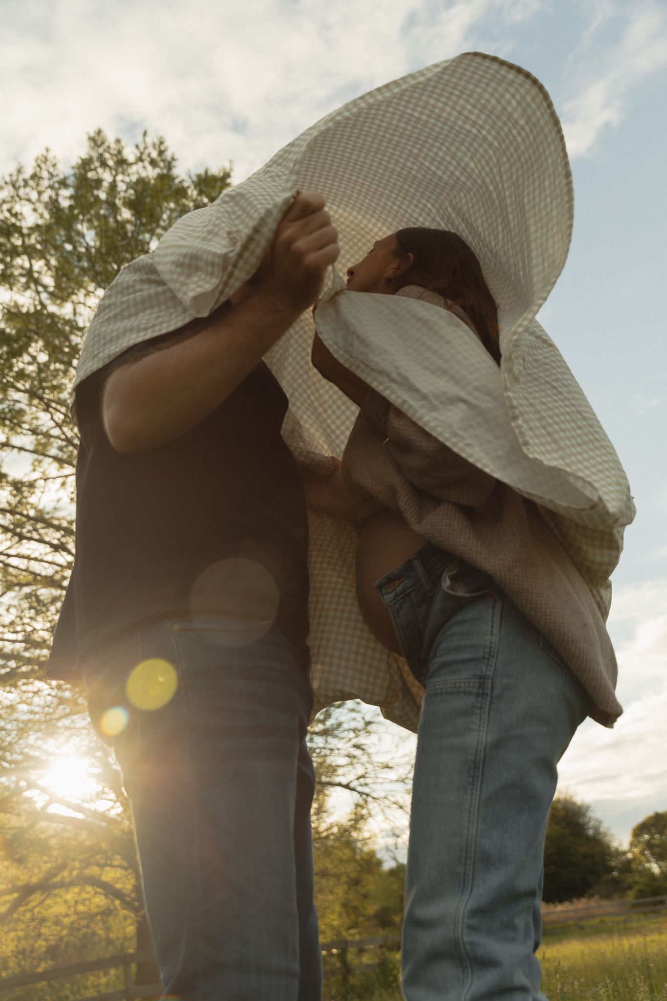 Two people sharing a blanket over their heads in a sunlit park, with lens flare and green trees in the background.